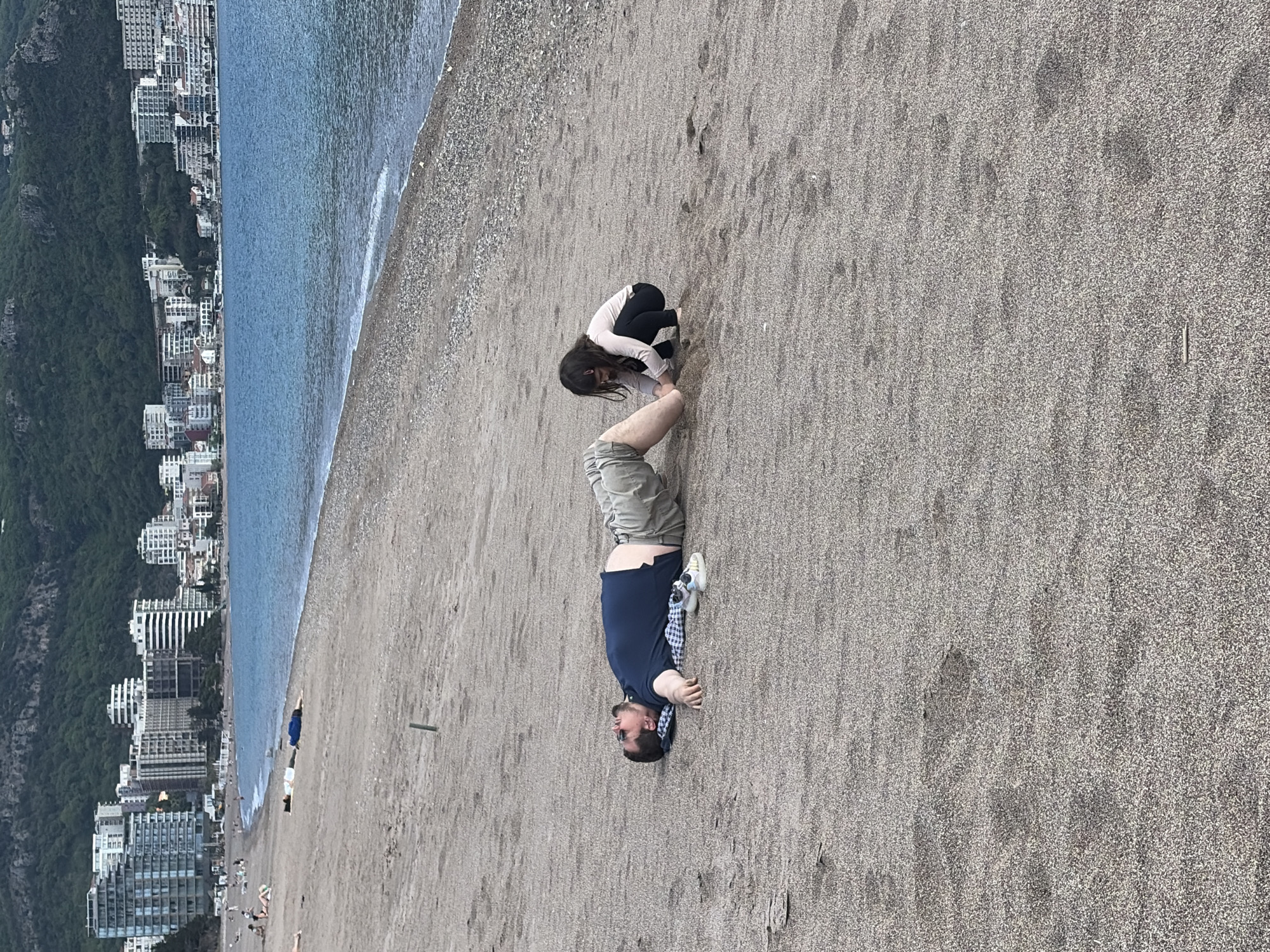 A man in a blue shirt and khaki shorts is lying on a beach, with a girl kneeling next to him. The beach is sandy and stretches along the coastline of the city of Boreti, Montenegro. A line of buildings and lush vegetation are visible in the background along a mountain. The sea borders the beach, and the sky is overcast.