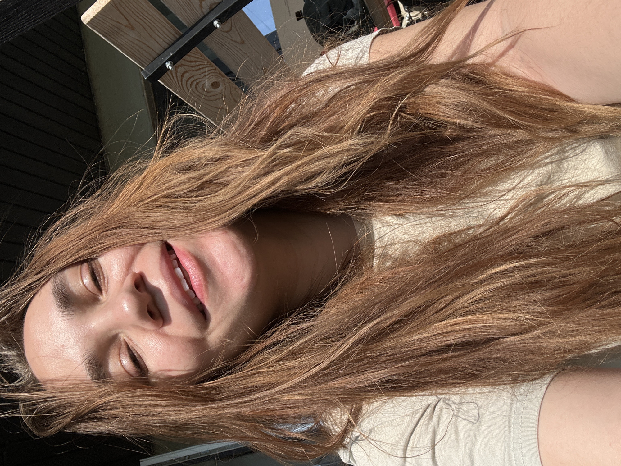 A close-up, rotated image depicts a person with long, light brown hair. The person's eyes are closed, and they have a slight smile revealing their upper teeth. The skin appears light-toned and reflects sunlight. The person is wearing a light-colored shirt. The background is partially obscured by the hair and includes elements of wooden and metal structures. There is no location information present in the provided data.