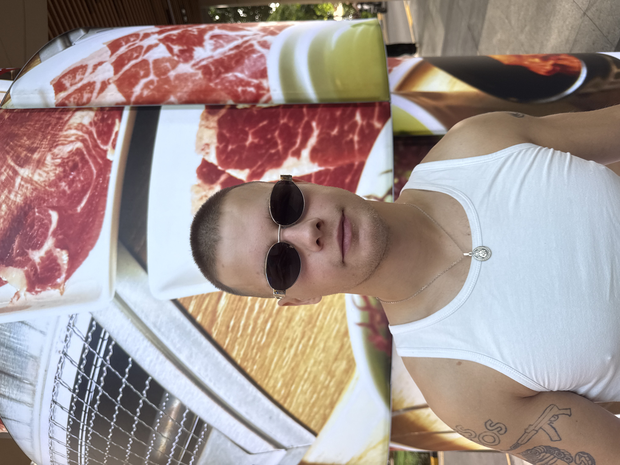 A young man with short hair and sunglasses stands in front of a large cylindrical structure covered in images of raw meat. He is wearing a white tank top and a silver necklace with a pendant. Tattoos are visible on his left arm, including what appears to be a firearm. The background shows a blurred street scene with trees and a streetlamp. The cylindrical structure is likely a part of an advertisement for a restaurant or food establishment. The scene is set outdoors, possibly on a sidewalk or plaza.