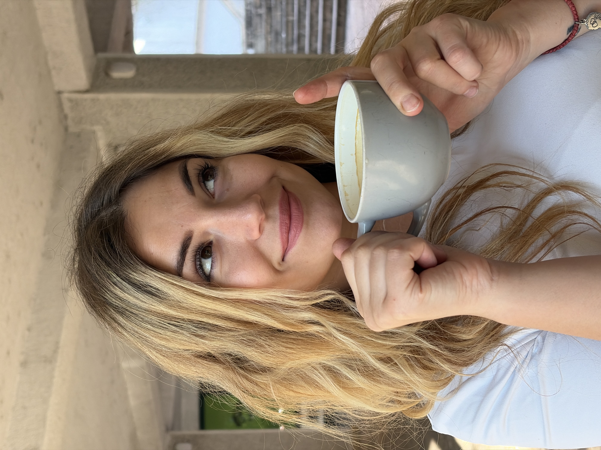A close-up, portrait-style image shows a young woman with long, blonde hair, holding a light grey mug with a frothy drink. She is looking upwards and slightly to her left with a gentle, contented smile. Her lips are colored with a matte pink lipstick, and her eyelashes are dark and defined. Her skin appears fair with a subtle blush on her cheeks.
The woman is wearing a light blue garment, possibly a t-shirt or blouse. A red bracelet with a paw print charm is visible on her left wrist. Her hands are positioned around the mug, with one hand holding the handle and the other gently resting on the side.
The background suggests an outdoor setting with architectural elements. Light-colored, textured columns or walls are visible on the left, with a hint of a staircase or elevated walkway in the distance under a bright sky. The lighting indicates it is daytime, likely sunny, creating soft shadows. The overall mood is relaxed and pleasant. No visible text is present in the image. The location context is Budva, Montenegro.