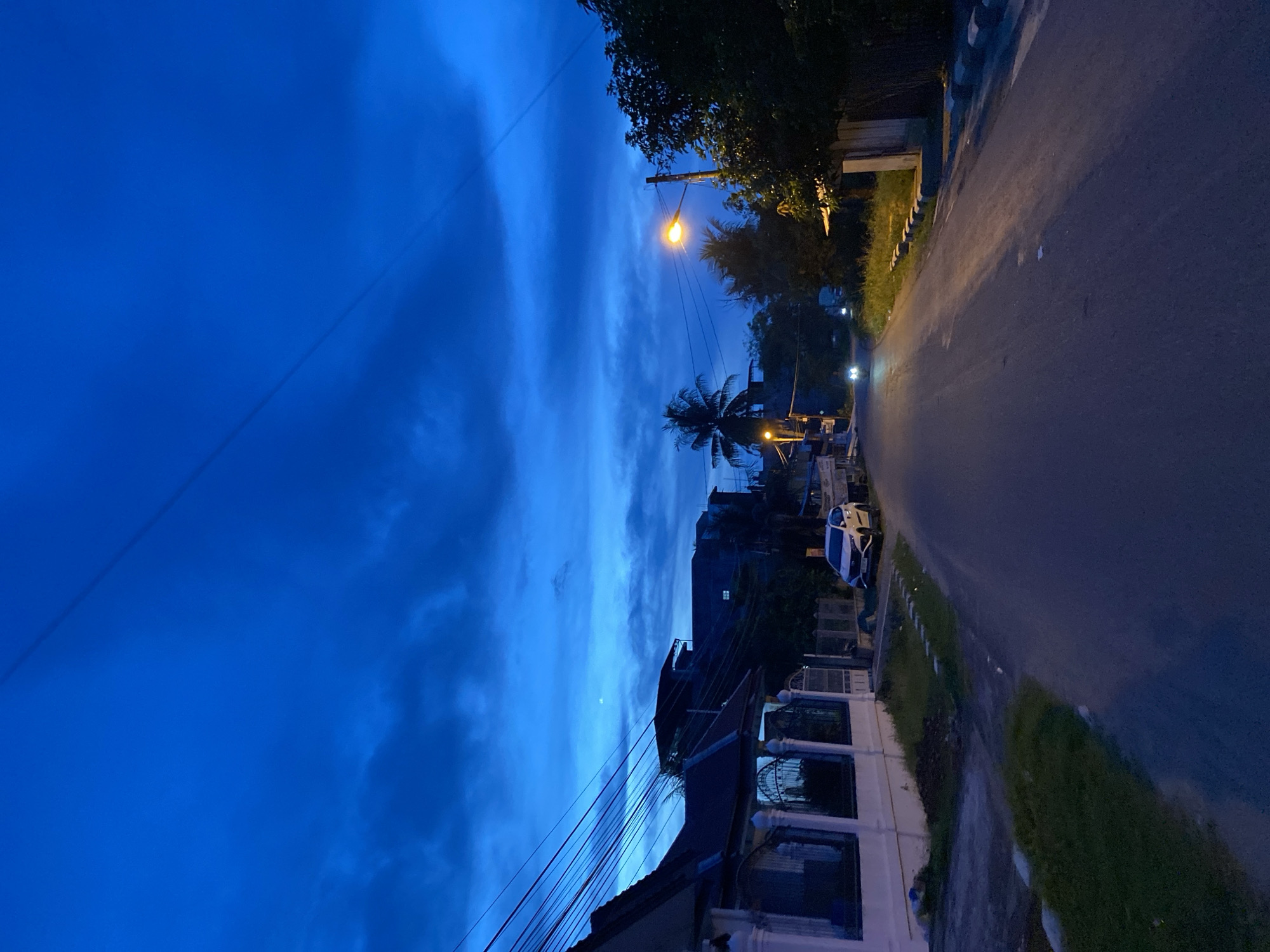 The image shows a street in Lhokseumawe, Indonesia, at dusk or dawn. The sky is a deep blue with some clouds, suggesting it is either twilight or predawn. There are several houses on the left side of the street, one with a white fence and decorative pillars. A white car is parked on the right side of the street. Further down the street, a streetlight is illuminated, and the headlights of an approaching vehicle are visible. Trees and foliage are present along the sides of the road. Overhead, power lines are strung across the scene. The overall atmosphere is quiet and serene, capturing a transitional moment of the day in a residential area.