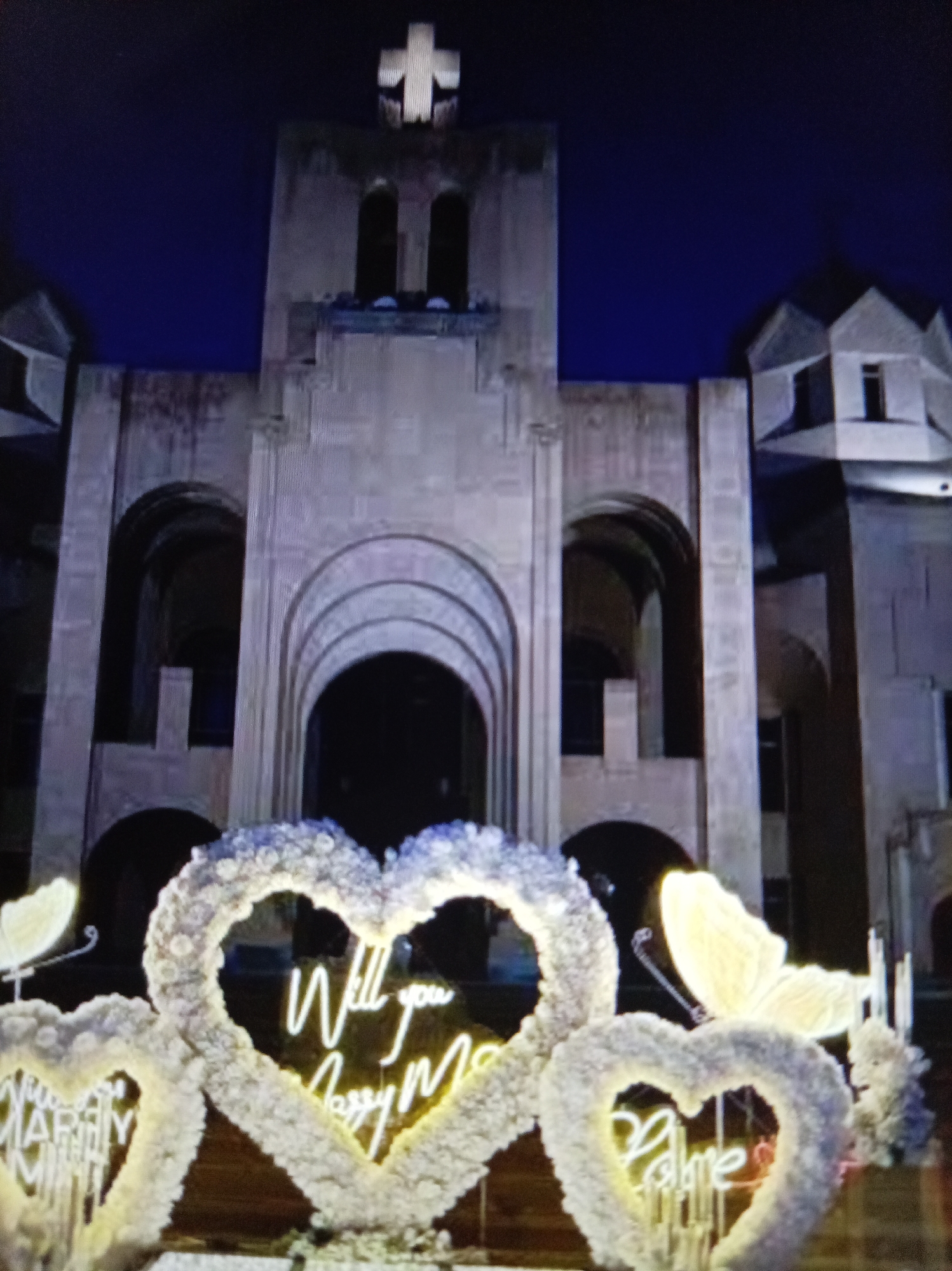In an exterior nighttime shot, a large, stone building with architectural features resembling a church or cathedral is illuminated against a dark blue sky. A prominent white cross tops the central structure. In the foreground, three large heart-shaped floral arrangements are displayed. The left heart, partially visible, has neon lettering that appears to read "MARRY ME." The central heart features neon text spelling out "Will you Marry Me." The right heart has neon text that reads "Love." Large, illuminated butterfly decorations are positioned near the floral hearts. The building's facade features arched entrances and windows. The scene suggests a romantic proposal or celebration.