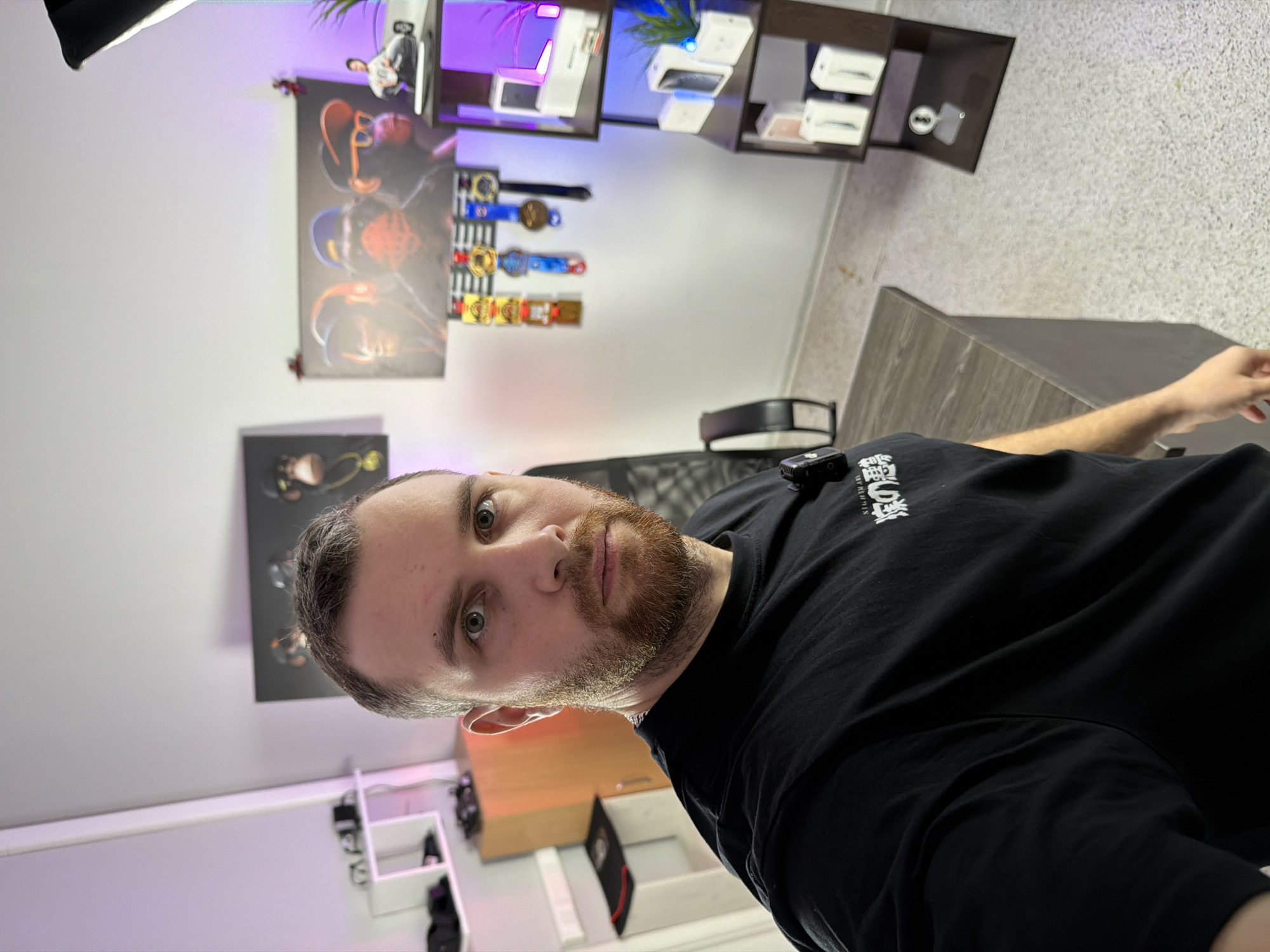 The image depicts an indoor scene, specifically a room in the city of Unknown, Unknown. A light-skinned male, approximately 25-35 years old, with short light brown hair, a full reddish-brown beard, and light-colored eyes, occupies the foreground. He is wearing a black t-shirt with white text, partially visible as "NIGHTMARE" and Japanese characters. He is looking directly at the camera, capturing a selfie-style photograph.
The room has light grey walls and a speckled light-colored floor. Purple LED strip lighting illuminates the upper perimeter of the room, creating a distinct glow. A softbox light source is visible in the upper right.
To the left, a white floating shelf holds various electronic items and cables. Below it, a two-door wooden cabinet is visible, with a lower white shelf holding a black object. Two framed artworks are mounted on the left wall: one depicting three abstract monkey-like figures, and another displaying a golden necklace in a black frame.
In the background, a large framed artwork on the rear wall portrays three stylized figures reminiscent of "Bored Ape Yacht Club" art, illuminated by purple light. Below this artwork, a black rack displays multiple medals of varying finishes (gold, silver, bronze) on colorful ribbons. To the right, a dark brown, multi-tiered display unit showcases several white smartphone boxes on its shelves. The top shelf of this unit also holds a small figurine and a plant, illuminated by purple light. The middle shelf contains more smartphone boxes and another small plant with blue illumination. The bottom shelf presents additional smartphone boxes and a transparent display stand. A black office chair with a mesh back is partially visible behind the subject. A dark grey desk with a wood-grain pattern extends from the left foreground towards the center.
