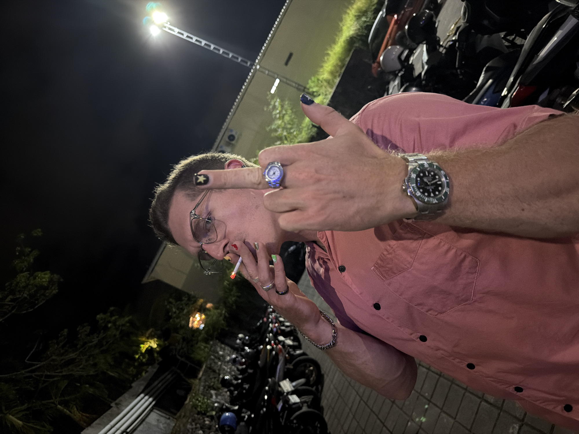 A person is shown in this low-angle outdoor night-time shot, standing in what appears to be a parking lot filled with motorcycles. The person is wearing glasses and a coral-colored button-up shirt. They are smoking a cigarette and holding up their middle finger, which is decorated with a black star design on the fingernail. The person is also wearing rings on their fingers and a silver wristwatch on their left wrist. The background shows a line of parked motorcycles, some vegetation, and a lit pole with a light fixture at the top. The ground is paved with interlocking tiles. The location is identified as Kuta Selatan, Indonesia.