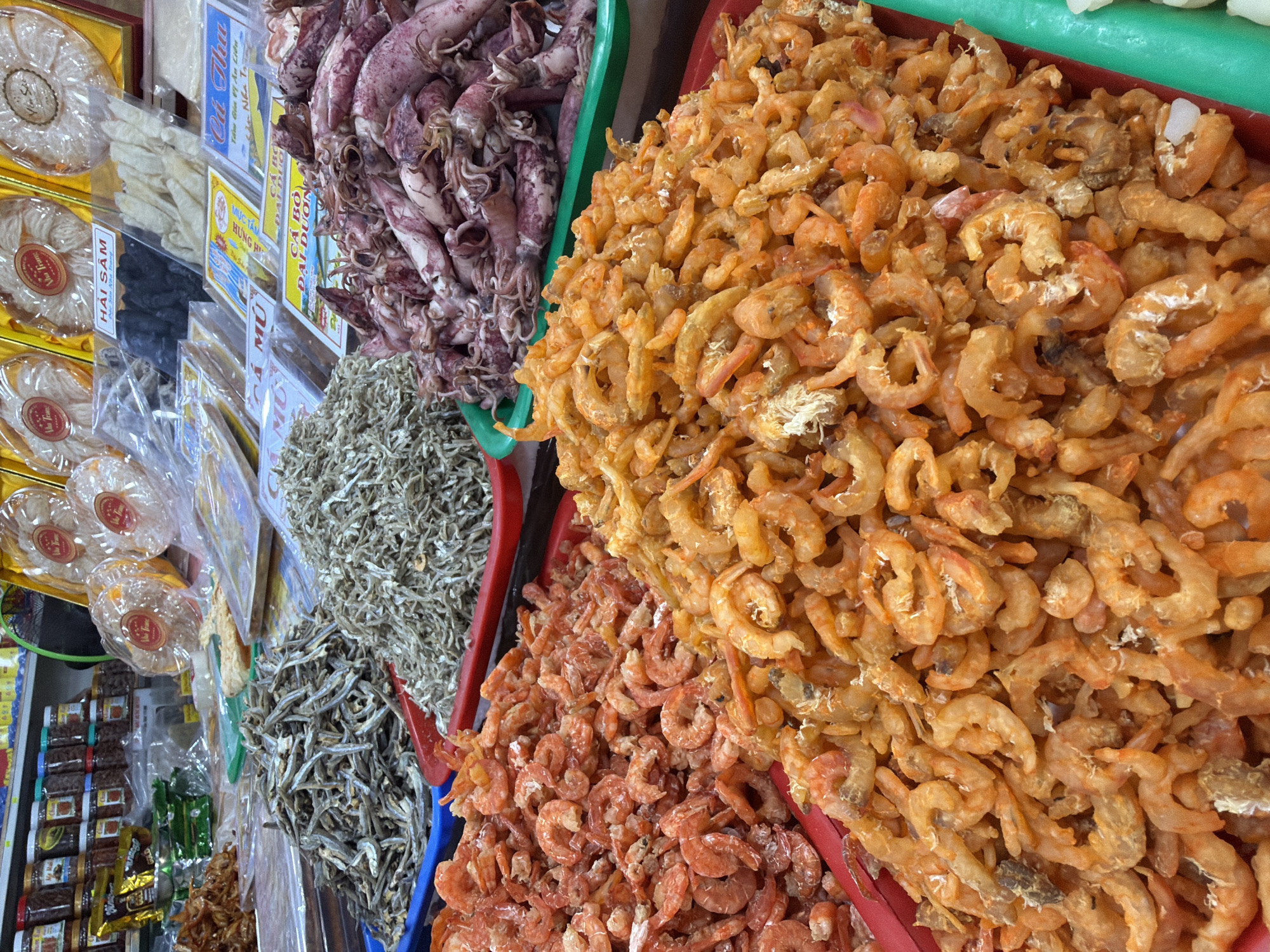 This is a top-down, eye-level, close-up shot of a food stall in Nha Trang, Vietnam, likely a market or shop. The foreground is dominated by large piles of dried seafood, primarily small, whole dried shrimp in various shades of orange and pink. To the left, a tray of larger, more prominently orange dried shrimp is visible. Behind these, to the left and center, are two large piles of small, silvery-grey dried fish, possibly anchovies. In the upper right corner, a container of dried squid, ranging in color from pale pink to deep purple, is present. The seafood is displayed in red and green plastic trays. In the background, shelves are stocked with jars of preserved foods and packaged goods. Signs are visible above the seafood, with Vietnamese text and imagery. The scene appears to be well-lit, with the focus on the abundance and variety of dried seafood.