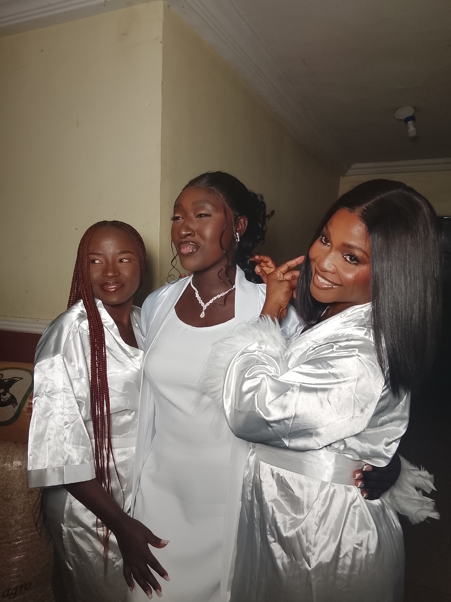 Three women are indoors, wearing white. The woman in the center wears a white dress and a white shawl, along with a necklace and earrings. The other two women wear white, shiny robes. The woman on the right smiles at the camera, while the woman on the left looks slightly off to the side. The location is the city of Unknown, Unknown.