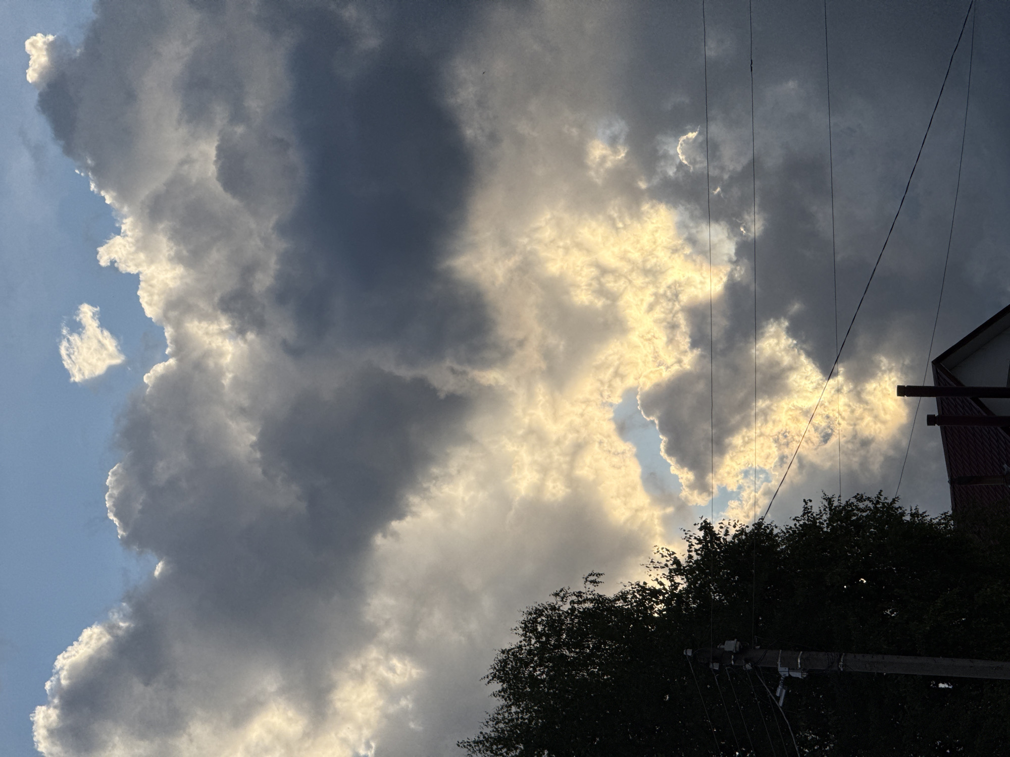 The image is a low-angle shot of a dramatic sky filled with an assortment of clouds, suggesting a transition in weather. The clouds are a mix of dark, ominous masses and bright, illuminated formations where sunlight is breaking through. The lighting creates a strong contrast, with pockets of brilliant yellow and white light piercing through the darker gray and blue hues of the sky.
In the lower right and lower central parts of the frame, the dark silhouette of a dense tree canopy is visible, with its leaves obscuring what lies beneath. Rising from behind the trees on the right edge of the image is the corner of a building with a red roof and dark brown walls, topped with a lighter trim. Multiple power lines stretch across the sky from left to right, adding a man-made element to the natural scene. These lines are taut and appear to be suspended from utility poles, one of which is partially visible in the lower portion of the image.
The scene evokes a sense of impending change, perhaps a storm brewing or passing. The time of day is likely late afternoon or early evening, given the angle of the sunlight and the dramatic cloud formations. The weather appears to be mixed, with both sunshine and dark clouds present. The location context provided suggests this is in Vetluzhsky, Russia. There is no visible text in the image.