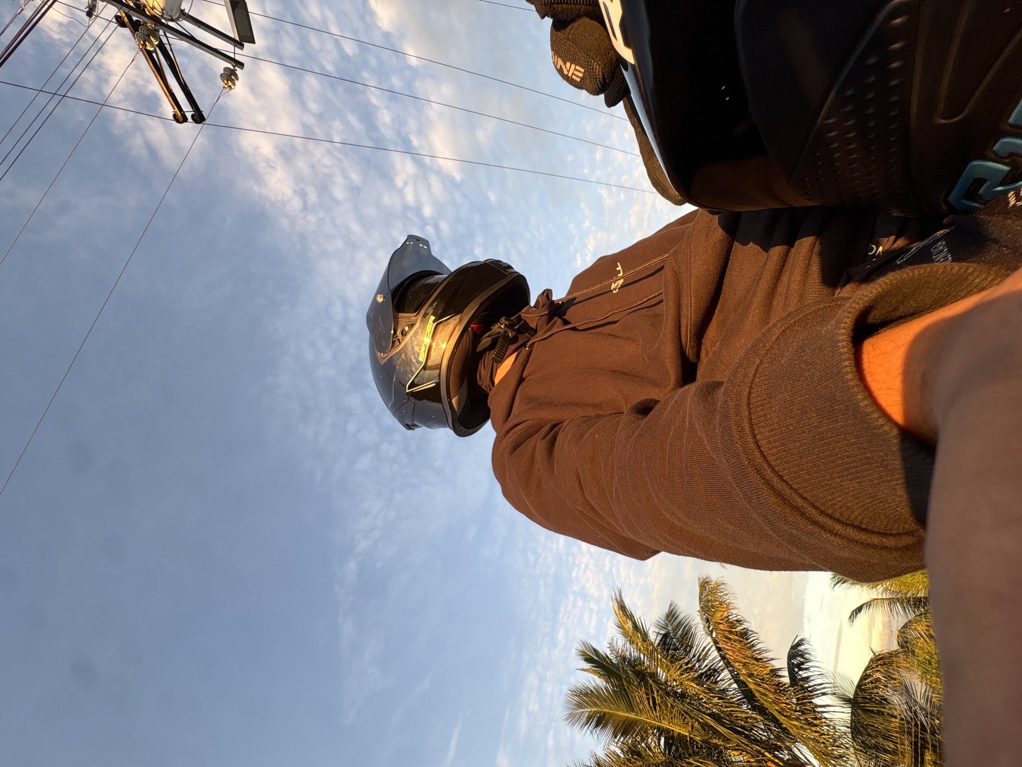 The image depicts a first-person perspective of an individual wearing a dark motorcycle helmet and a brown hooded sweatshirt, seemingly seated on a motorcycle. The individual is viewed from a low angle, looking upwards towards a bright, cloudy sky. To the left, palm trees are visible, suggesting a tropical environment. In the upper right portion of the image, power lines and utility poles are present. The lighting indicates it is either sunrise or sunset, casting a warm glow. The scene appears to be an outdoor setting, potentially along a road or in a more open area, consistent with locations within Davao City, Philippines, where such a combination of elements like palm trees, power lines, and open skies is common. No specific city landmarks are identifiable, but the overall environment aligns with a tropical urban or suburban landscape in Davao City.