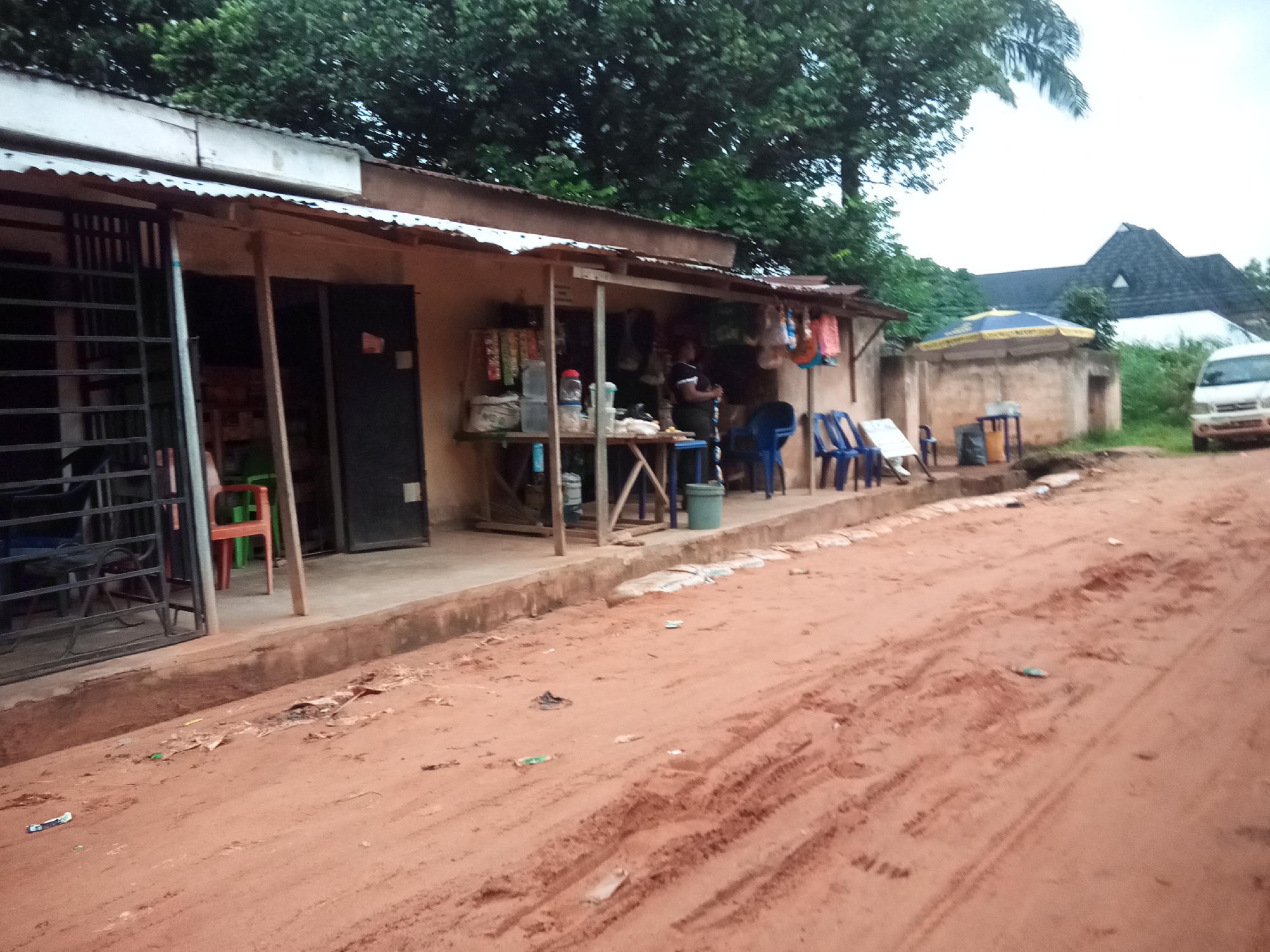 A roadside stall and its surroundings are depicted in Nawfija, Nigeria. In the foreground, a dirt road with visible tire tracks dominates the lower half of the image, littered with small pieces of debris. To the left, a simple, open-fronted stall with a corrugated metal roof is visible. It is constructed from wood and concrete block walls. Inside, various items are displayed on shelves, including what appear to be bottles of water, packaged goods, and bags. A person stands behind the counter. In front of the stall, several blue plastic chairs are arranged. Adjacent to the stall, a small, weathered structure with a blue and yellow umbrella stands on a concrete base. Further to the right, a white van is parked on the side of the road. In the background, lush green trees form a dense canopy, and a building with a dark, steeply pitched roof is partially visible. The sky is overcast, suggesting a cloudy day.