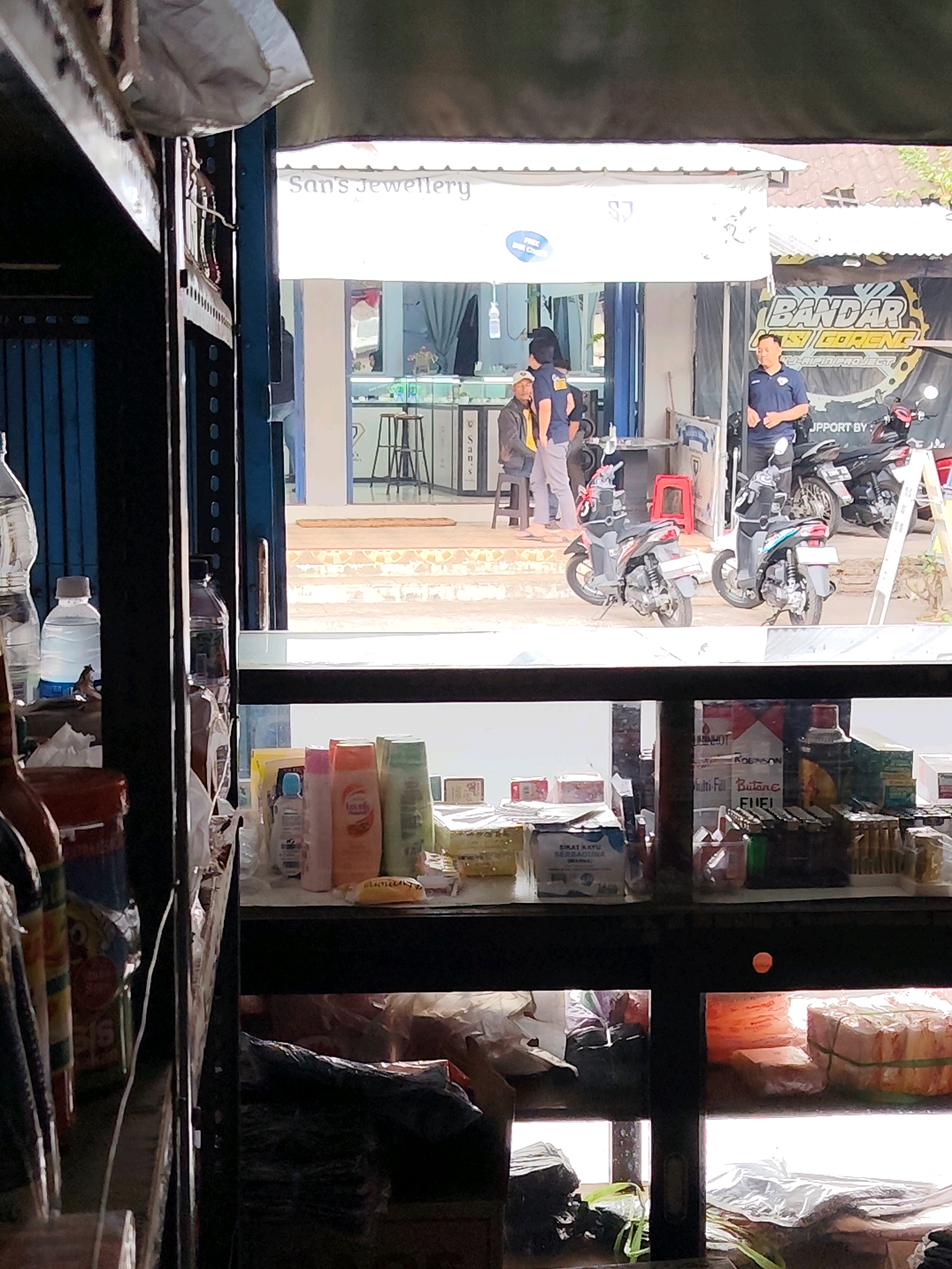 The image captures a street-level view of a storefront in Dampit, Indonesia, as observed through an open window. On the left, a shelf displays various bottles, including a large clear bottle with a white label and a blue cap, and a smaller clear bottle with a white label and grey cap, both appearing to be water bottles. To the right of these, on a lower shelf, a collection of toiletries is arranged, featuring several bottles of shampoo or body wash in various colors and brands, including one with an orange label reading "Love" and another with a light green label. A small blue bottle is also visible in this grouping.
The central part of the image reveals the exterior of a shop with signage that reads "San's Jewellery". In front of the shop, two individuals are visible. One, wearing a dark blue shirt with a logo and light-colored pants, stands near a motorcycle. Another person in a grey jacket and a white cap is also present, facing away from the viewer. Further into the shop, more display cases with items inside are visible, along with a stool.
To the right of the "San's Jewellery" signage, a larger banner is displayed with the text "BANDAR GORENG" in bold yellow lettering. This banner also features an image of a motorcycle and the words "RJU-AJA PROJECT". Two motorcycles are parked in front of this area, and a man in a dark blue shirt with a logo is standing near them. A red stool is placed to the left of the motorcycles, inside the shop's doorway. The background shows a building with a tiled roof.
The lower section of the image, viewed through another window, shows shelves filled with various items. On the left, stacks of what appear to be packaged goods, possibly food or household items, are present, some wrapped in plastic. Further to the right, more boxes and plastic-wrapped items are stacked. The overall scene suggests a commercial area with shops selling a range of products.