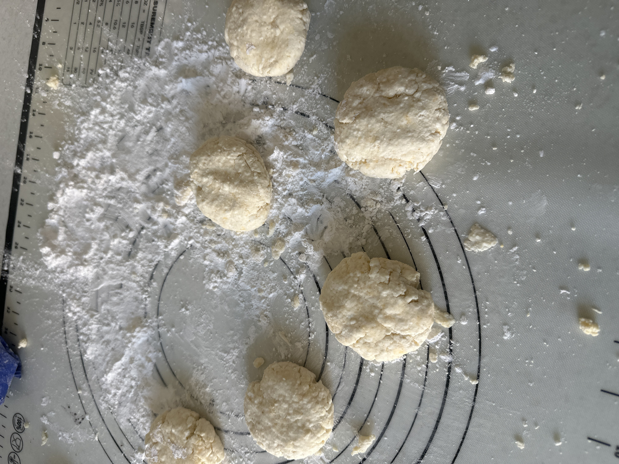 Seven balls of dough, each approximately three inches in diameter, are scattered across a white silicone baking mat. The mat has black concentric circles and ruler markings printed on it, suggesting it is a measuring mat. A dusting of white flour covers parts of the mat and the dough balls. The dough appears to be pale yellow and slightly lumpy. One corner of the mat has a blue cloth partially visible. The scene is a close-up shot, with no other environmental details provided beyond the mat itself. There are no people visible in the image. The location in the city of Boo, Sweden, is not discernible from the visual information provided in the image.