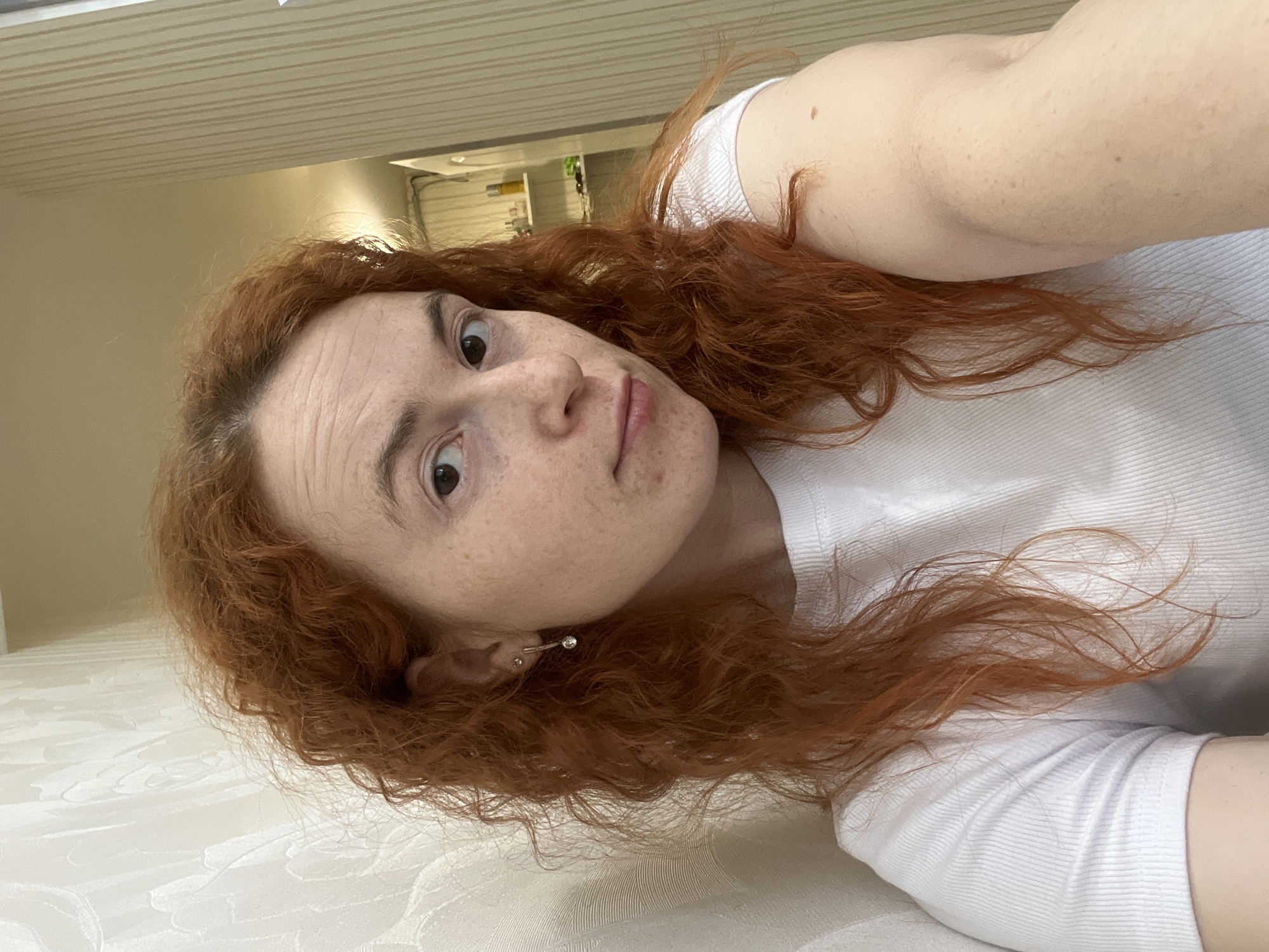 A woman with reddish-brown curly hair is lying on a white textured surface. She is wearing a white t-shirt. She has fair skin with freckles. She is wearing a silver earring in her left ear. The background appears to be an interior space with a beige wall and ceiling. A hallway with various objects is visible in the distance. There is no evidence suggesting that this photo was taken in Chaikovsky, Russia.