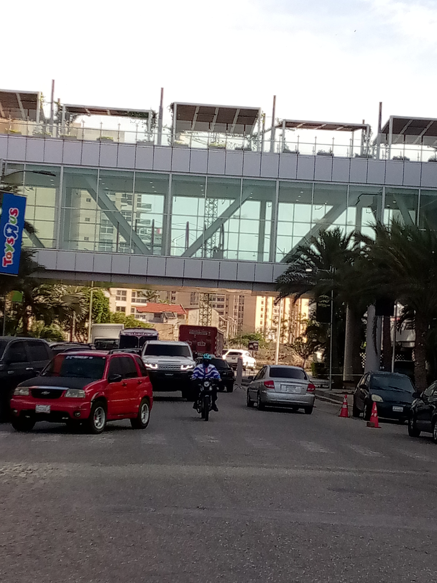 The image shows a street scene in Porlamar, Venezuela, with several cars, a motorcycle, and a pedestrian overpass. A red Chevrolet Tracker is in the foreground. A silver sedan is in the middle ground, followed by a dark-colored car next to two orange traffic cones. A white SUV is visible behind the Tracker, and a motorcyclist is riding in front of it. The Toys "R" Us sign is affixed to the side of the pedestrian overpass. The backdrop includes buildings and palm trees.