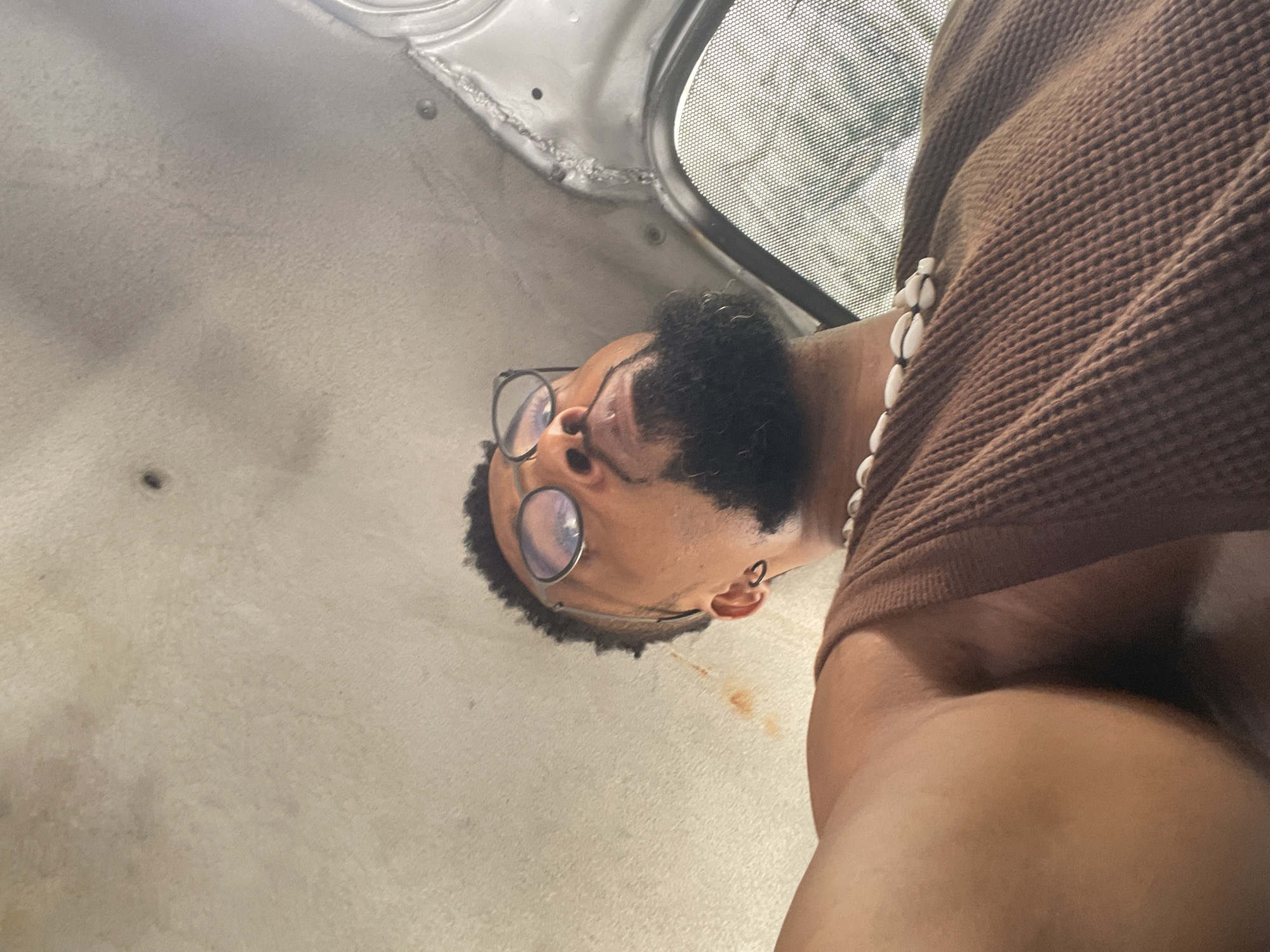 An intimate portrait of a person, likely male, positioned horizontally across the frame due to the image's orientation. The subject has dark skin, wears circular-framed glasses, and a short beard. A white cowrie shell necklace is visible around their neck, and they are dressed in a brown, textured, long-sleeved top. The shot is taken from a low angle, looking upwards.
The setting appears to be inside a vehicle, with a light-colored, possibly off-white or grey, ceiling or panel filling most of the background. Part of a window or sunroof with a distinct grid pattern, possibly a mesh screen, is visible in the upper right. A small, reddish-brown stain mars the light-colored surface near the person's head.
The lighting is soft and natural, indicating daytime. The person's gaze is directed towards the top of the frame. No discernible specific activity takes place beyond looking up, and the expression is neutral. There is no visible text. While the location is provided as Lagos, Nigeria, the image itself offers no direct visual cues to confirm this.