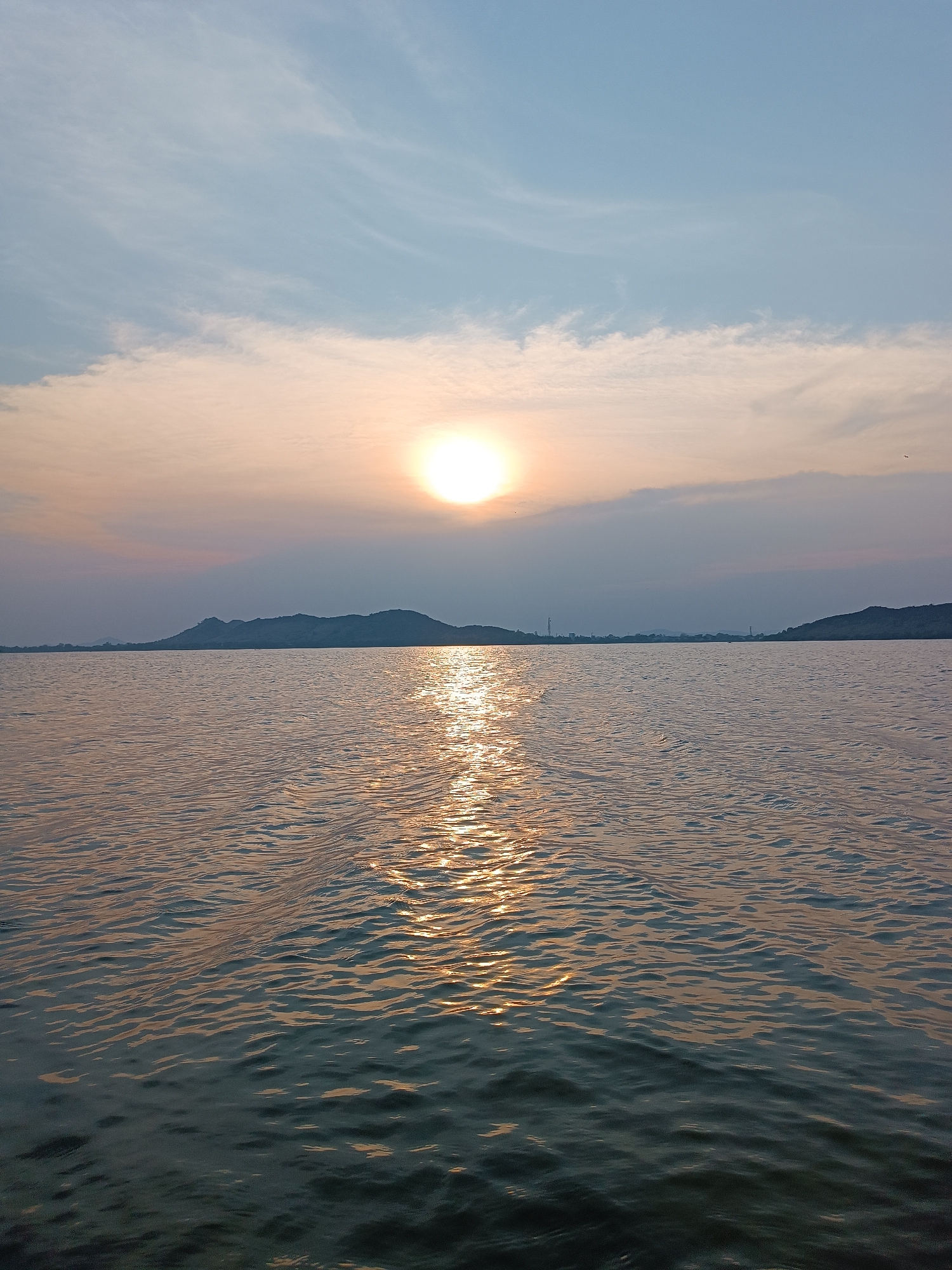 The image captures a wide expanse of water under a sunset sky. The sun, partially obscured by clouds, casts a bright reflection that shimmers across the water's surface. In the distance, silhouetted landmasses or hills are visible. The sky is a blend of light blue and soft orange hues, indicative of the setting sun. The water surface is slightly rippled, creating a textured pattern reflecting the sunlight. The image is taken in the city of Kammavaripalli, India.