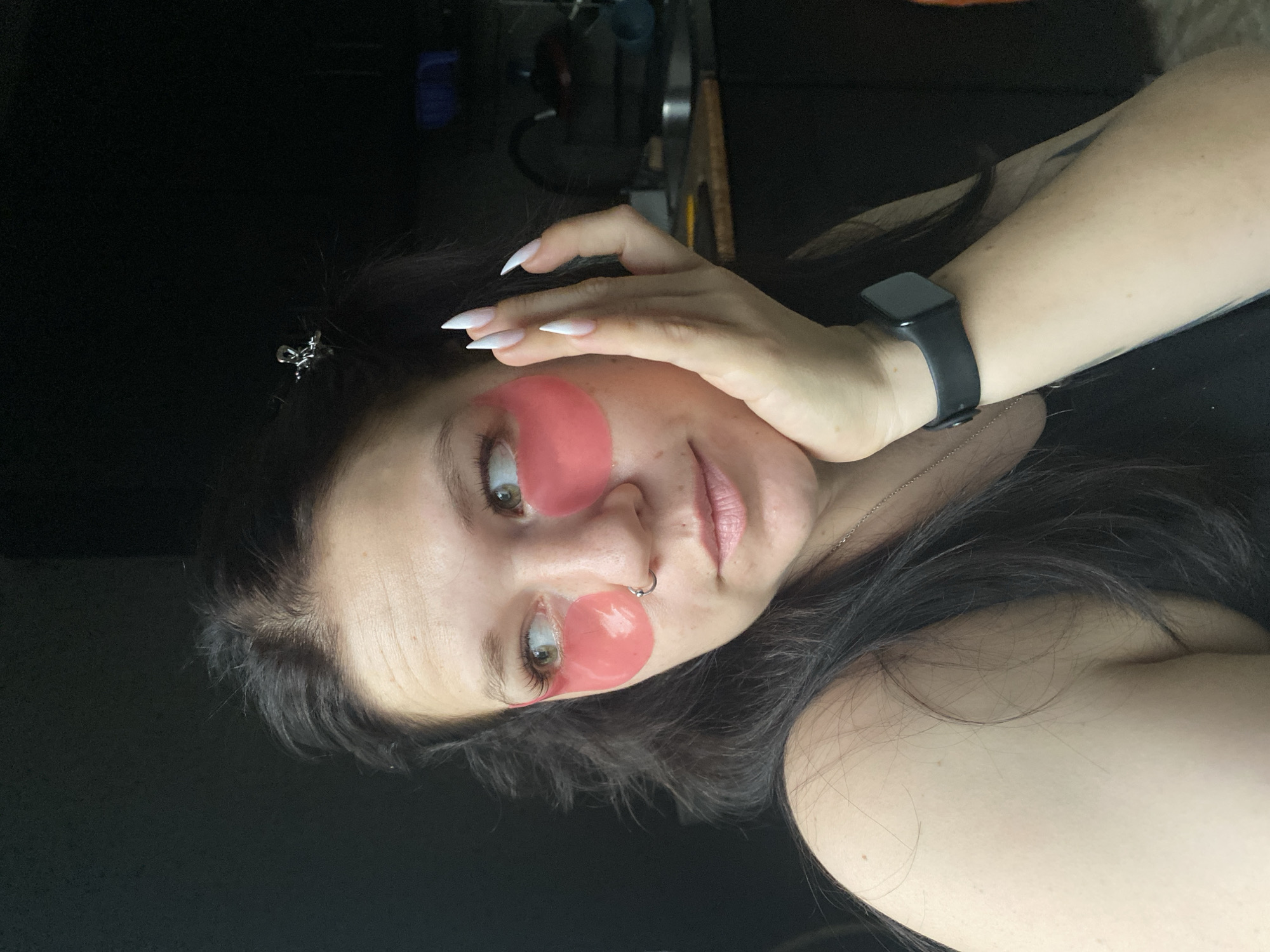 A woman is seen in a close-up shot, wearing pink under-eye patches. She has long, white-tipped nails and a black smartwatch on her wrist. The background is dark and blurry. There is no indication of the location being in the city of Chaikovsky, Russia.