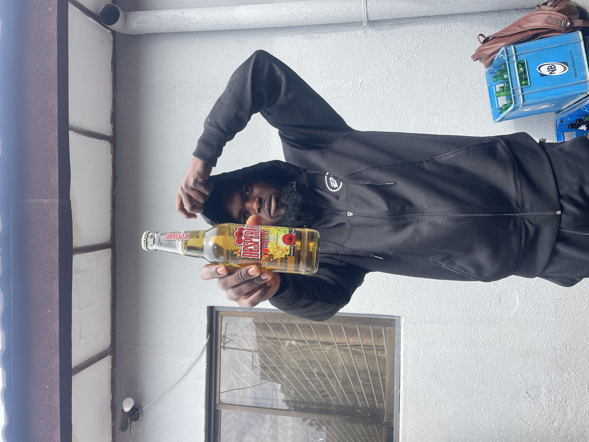 A man with dark skin and a beard, wearing a black hooded jacket with the hood up, poses for the camera. He extends his right arm forward, holding a clear glass bottle of Desperados Original Beer with tequila flavor. The bottle's label is visible, displaying "DESPERADOS ORIGINAL BEER," a sun or chili pepper logo, and "5.9% alc./vol." The man looks directly at the camera with a neutral expression.
The scene is set against a light-colored, textured wall, likely outdoors or in a semi-enclosed area like a patio. White pipes run along the top of the wall. To the left, a dark brown structural element is visible. Below the man's arm, there's a window fitted with security bars or a screen.
In the background, to the upper right, two blue plastic crates are stacked. The top crate contains several green glass bottles and displays an "NB" logo along with the text "Nigerian Breweries Plc," indicating its origin. A brown leather-like bag rests on top of the crates. The natural lighting suggests it is daytime. The presence of the Nigerian Breweries crate is a strong location cue aligning with the context of Jos, Nigeria.