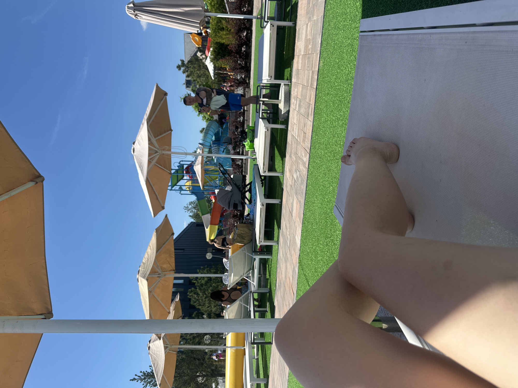 The image shows a sunny day at a water park in Khotianivka, Ukraine. In the foreground, a person's legs are stretched out on a lounge chair, with the artificial turf of the park visible in the background. The lounge chair is white with a light-colored fabric. Several other lounge chairs and umbrellas are arranged on the turf. Further back, people are seen relaxing or walking. Behind the lounge chairs, there are buildings and colorful water slides, indicative of a water park. The sky is clear and blue. A man is standing in the middle ground holding a baby.