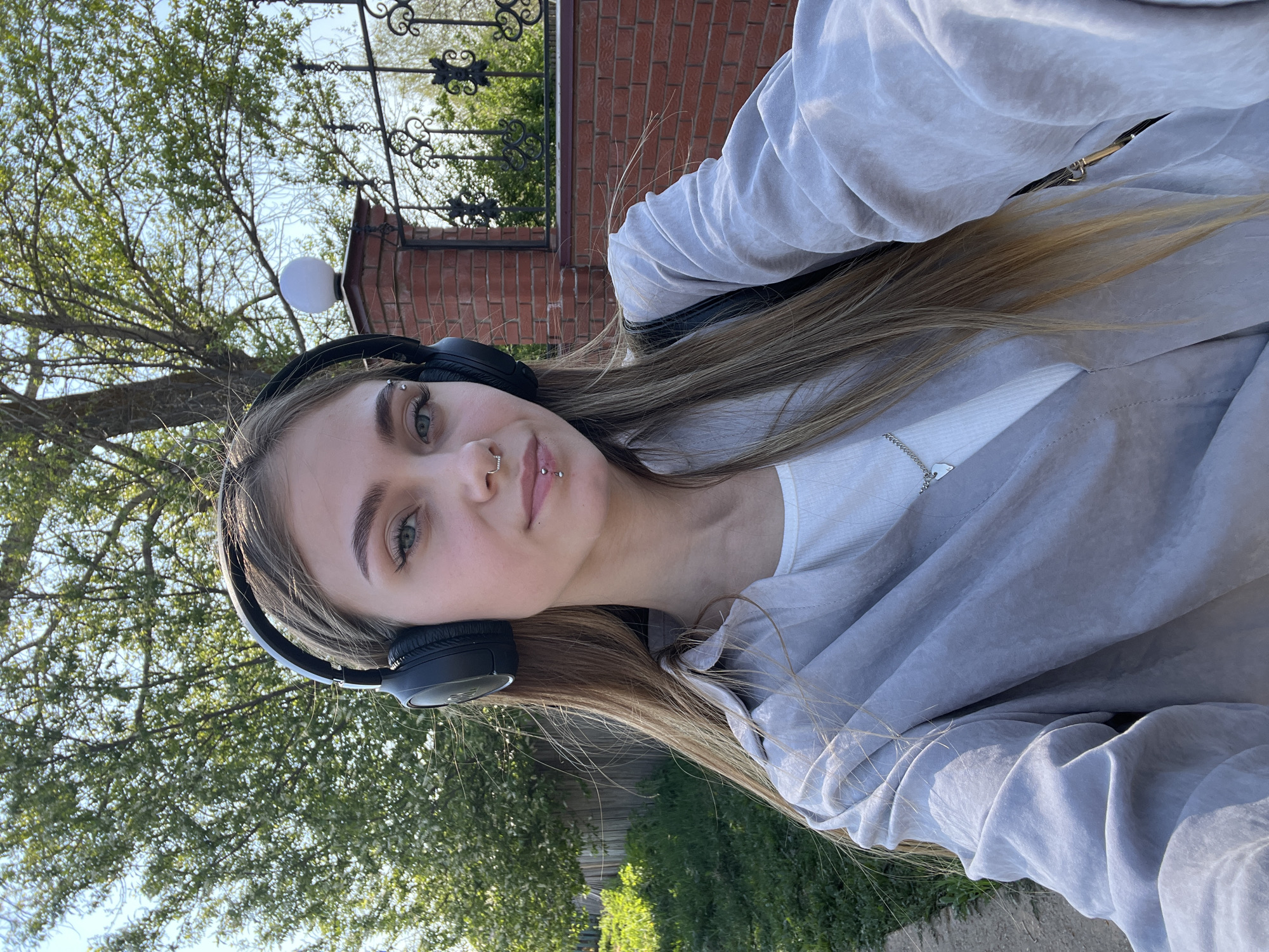 A young woman poses for a selfie outdoors. She has fair skin, light brown hair with blonde highlights, and green eyes. She has multiple piercings, including eyebrow, nose, and lip piercings. She is wearing black over-ear headphones, a white top, and a light gray zip-up jacket.
The background includes trees with green leaves, a brick wall, and a wrought iron fence. The location may be Фоки, Russia, though no features identifying the city are visible.