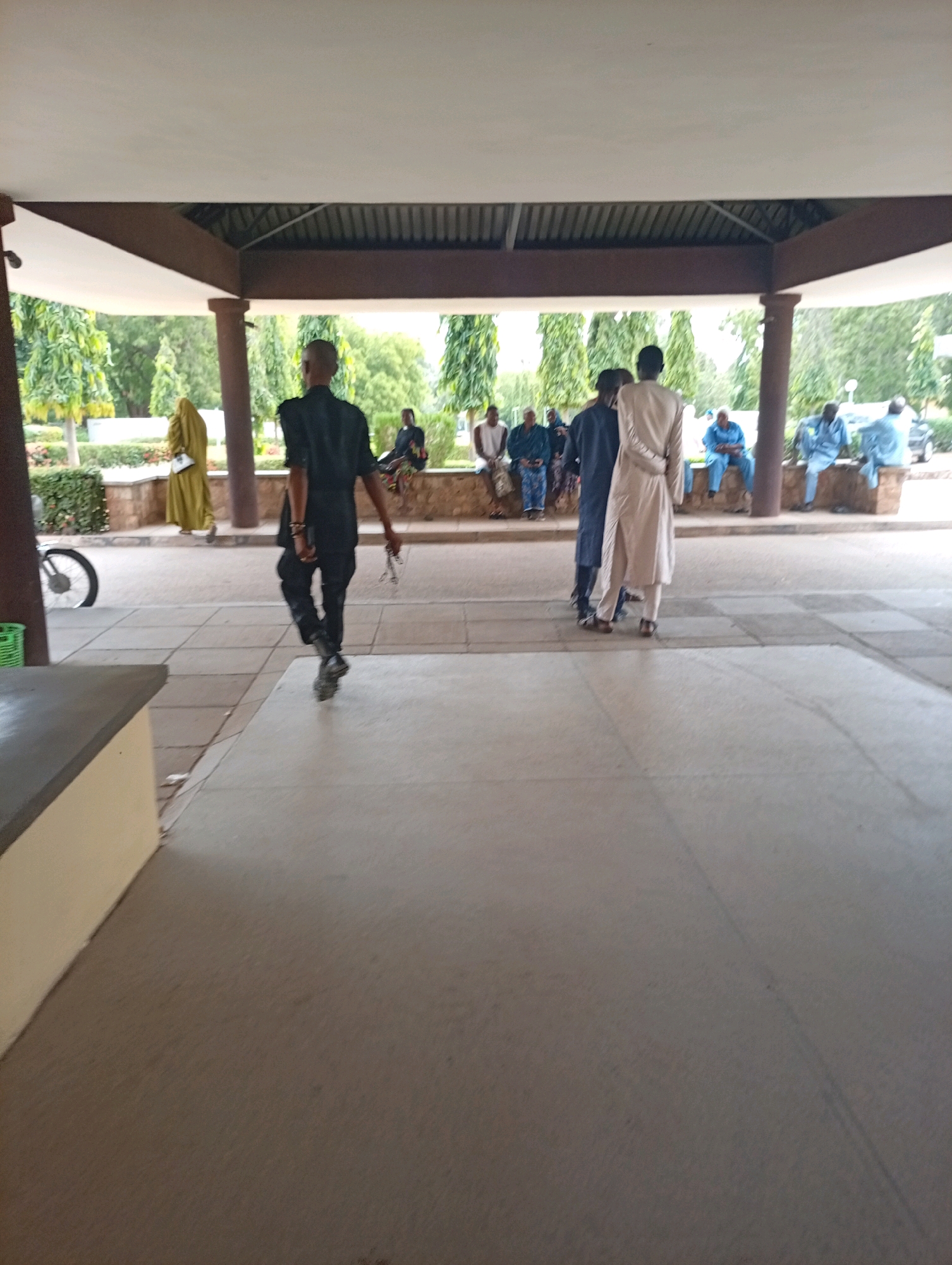 The image depicts an outdoor scene in Gombe, Nigeria, likely near an educational or institutional building given the presence of a covered walkway and manicured trees in the background. In the foreground, a man in a dark uniform, possibly security personnel, walks away from the camera. He is wearing dark trousers, a dark short-sleeved shirt, and dark shoes. He is holding a set of handcuffs in his right hand.
Further in the background, several individuals are seated on a raised ledge or bench under the covered walkway. These individuals are dressed in casual clothing, with some wearing light-colored robes and head coverings, indicative of local attire. One person in a yellow robe walks away from the camera towards the left. To the right, other individuals are also seated, many in light blue attire. A bicycle with a partially visible wheel is parked on the left side of the frame. The environment suggests a public space with a paved ground and a covered area for shelter. The overall atmosphere appears calm.