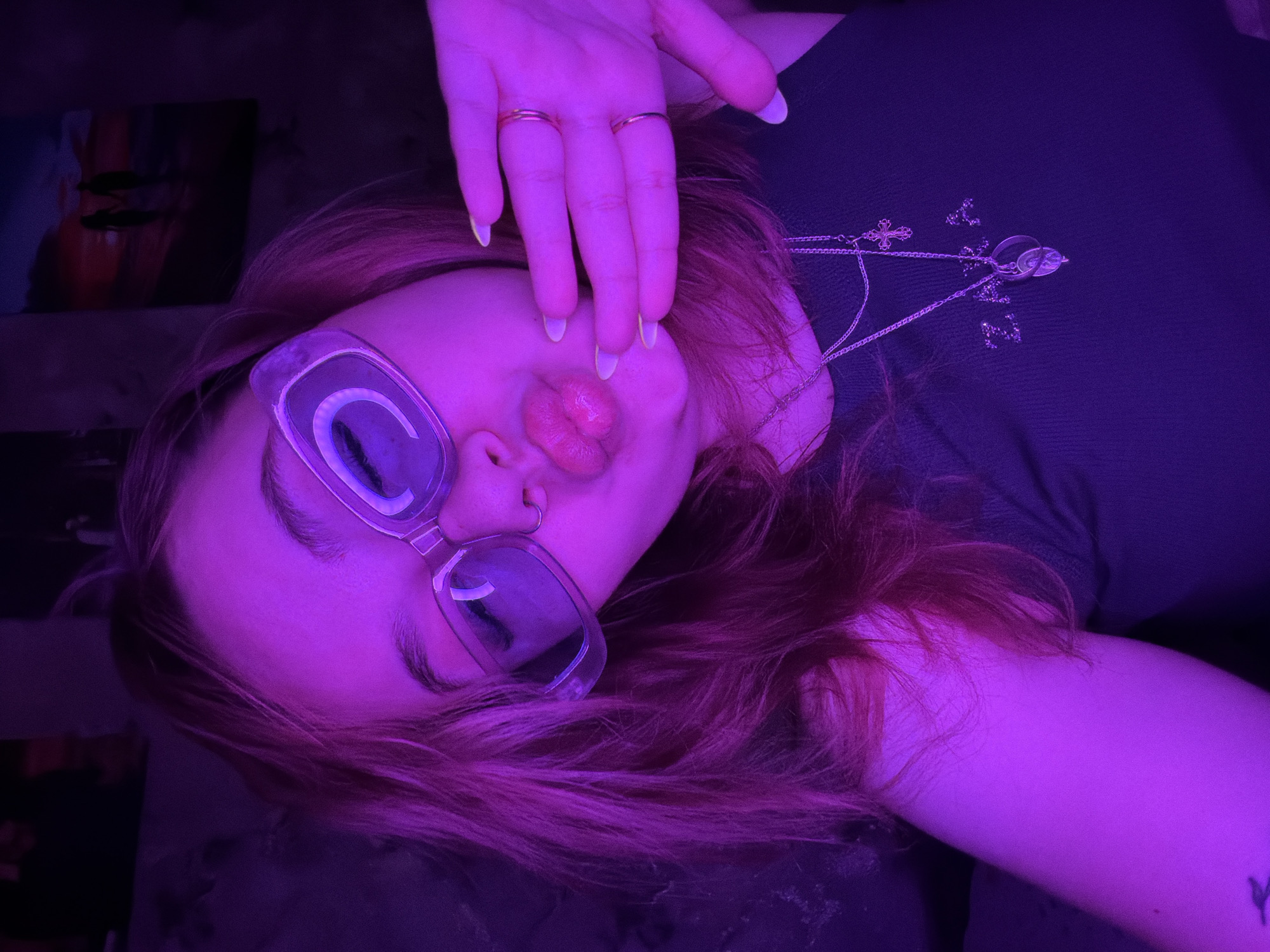 A person with reddish hair and glasses is lying down, facing upwards towards the camera. They have two rings on their fingers, and their hand is near their face. A necklace with multiple charms is visible on their chest. The lighting in the image is predominantly purple. The exact location in Chaikovsky, Russia, cannot be determined from this image alone.