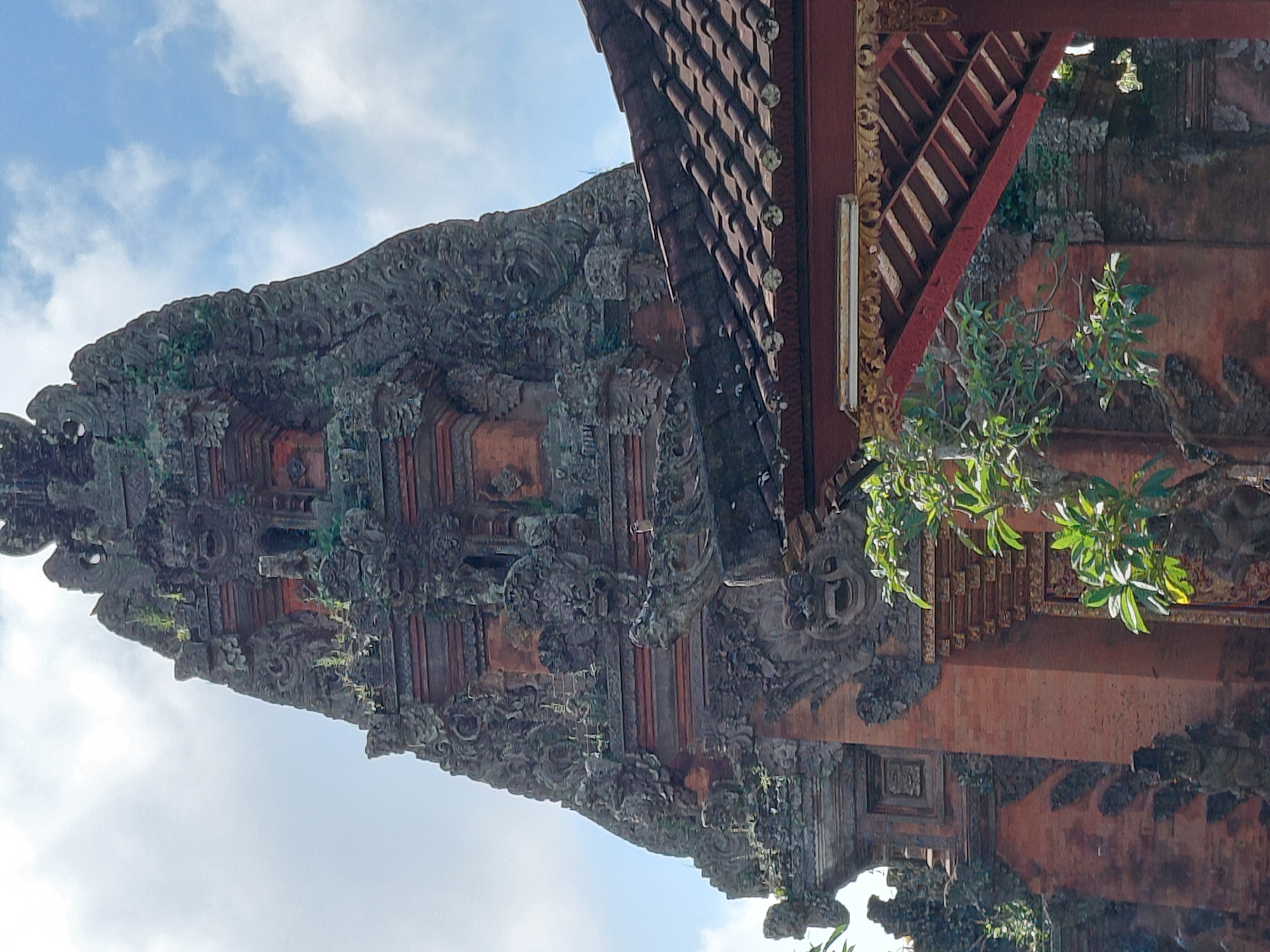 The image shows a close-up view of a Balinese temple structure, likely located in Ubud District, Indonesia. The temple features intricate stone carvings and red brickwork, with a towering, ornate structure dominating the upper portion of the frame. Lush green foliage and a tiled roof of a smaller adjacent structure are visible in the foreground and to the right, respectively. The sky above is partly cloudy.