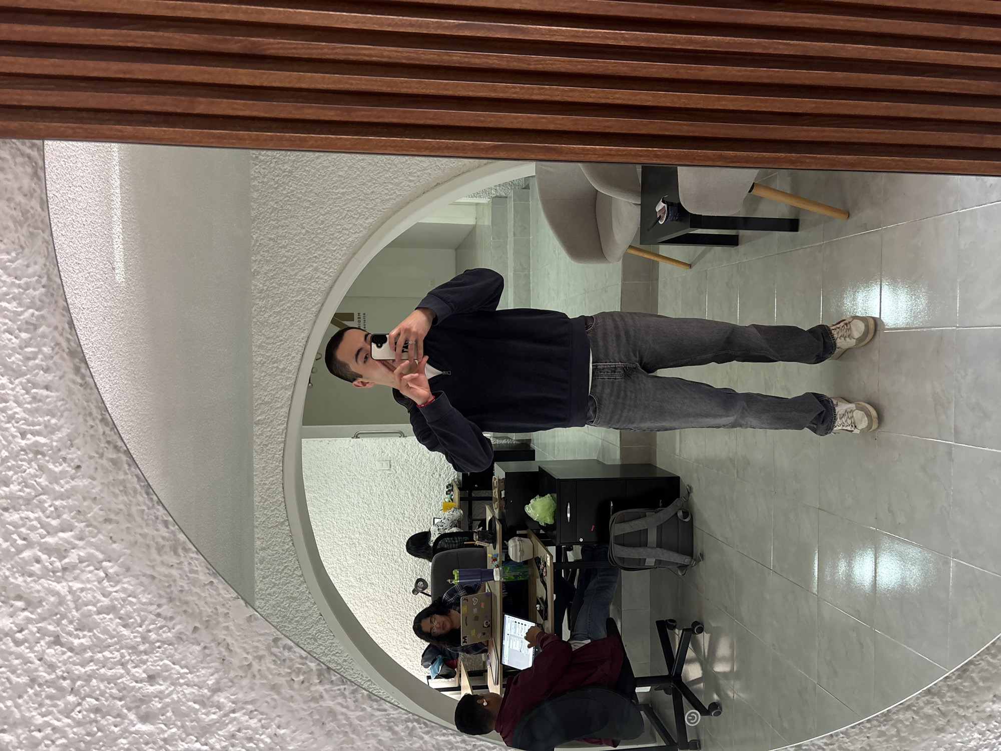 A man is reflected in an oval mirror in a co-working space in Puebla, Mexico. The man, facing the mirror, holds up a cell phone with a white case, apparently taking a photo of himself. He wears a navy blue sweatshirt, gray jeans, and white sneakers. The oval mirror has a decorative wooden border at the top and sits against a textured white wall. Behind the man in the mirror's reflection, are several people seated at desks working on laptops. The co-working space has white tiled floors and a light gray chair is visible in the background. There are black filing cabinets next to some of the desks.