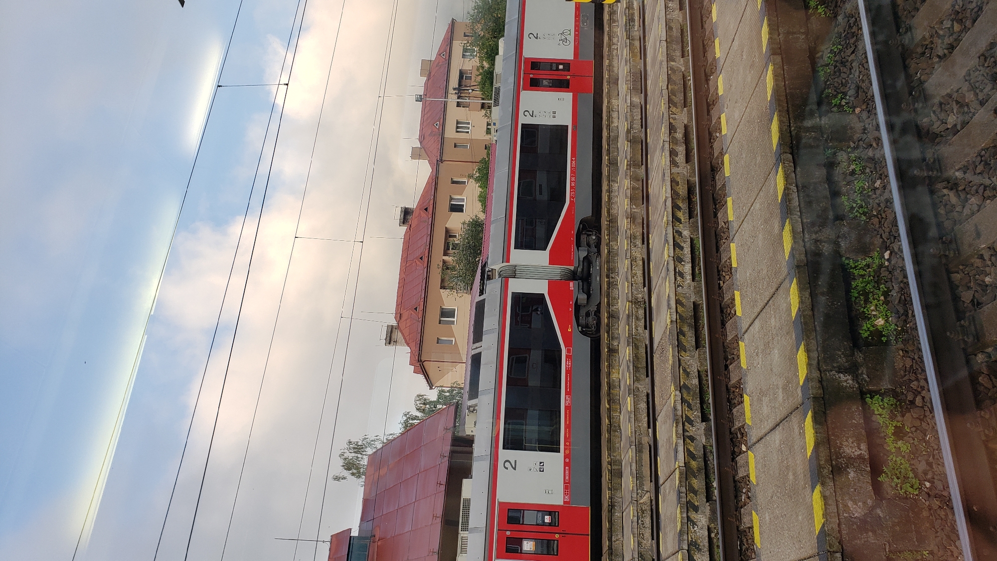The image shows a train at a train station in Margecany, Slovakia. The train is red and white and appears to be modern. The train station has multiple tracks and platforms, and there are buildings in the background. The sky is cloudy. There are no people visible in the image.