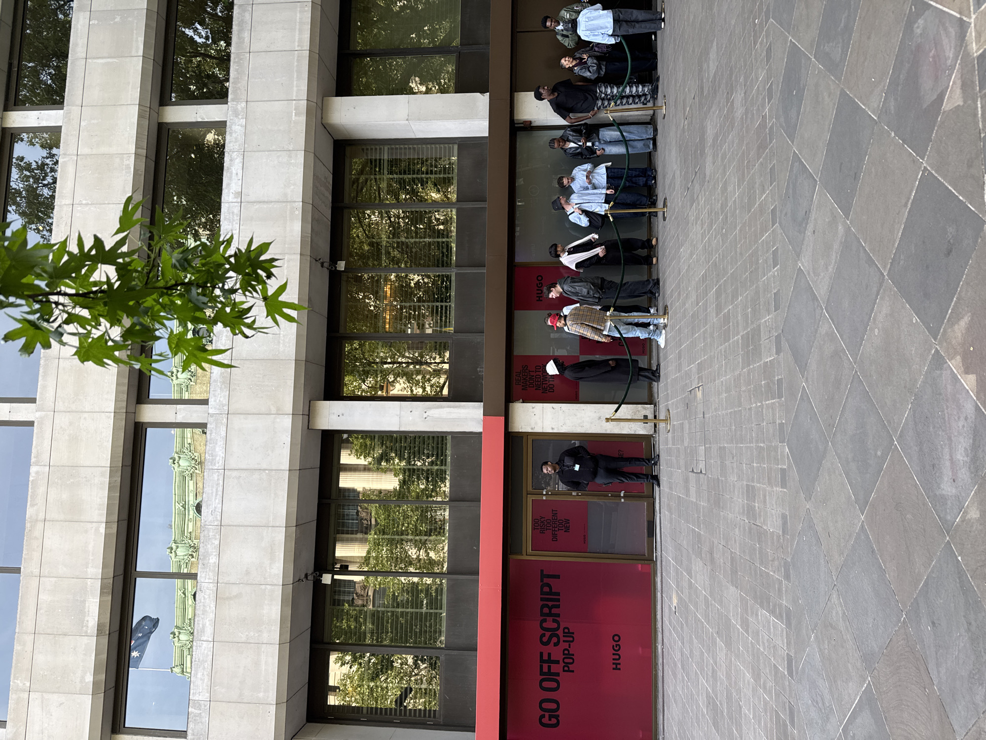 The image shows a queue of people outside a HUGO "GO OFF SCRIPT POP-UP" store. The store has a red facade with white lettering. A security guard stands by the entrance. A line of people stand in front of the store, behind a green rope line. The building is a multi-story structure with large windows. The windows reflect the Australian flag, suggesting the location may be Australia House in London. The pavement is made of grey stone tiles arranged in a diamond pattern. There are trees in the foreground.