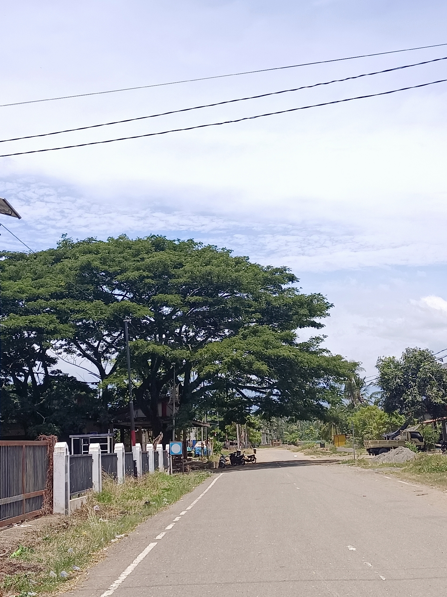 The image depicts a roadside scene in Meunasah Pupu, Indonesia. A paved road runs from the foreground into the distance, bordered on the left by a metal fence and a low concrete wall with vertical white posts. Beyond the fence, there are structures and vegetation. On the right side of the road, there is a dirt shoulder with grass, a pile of gravel, and some trees. In the background, a large, leafy tree dominates the left side of the frame, with smaller trees and buildings visible behind it. The sky is partly cloudy. Several motorcycles are parked on the side of the road near the large tree, and a truck is visible further down the road on the right. Power lines stretch across the top of the image.