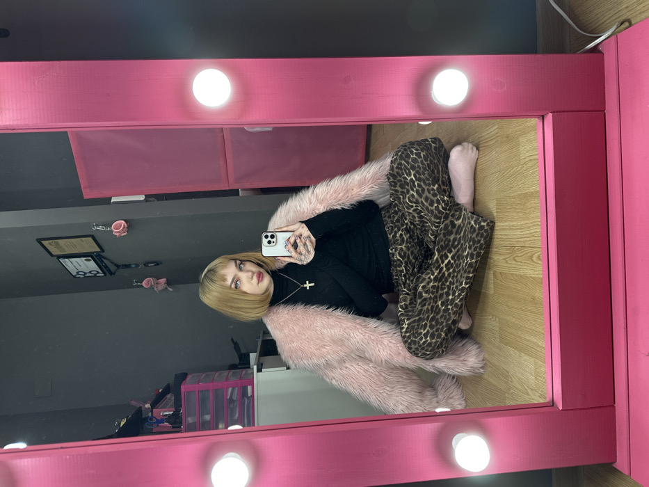 A blonde woman with bangs takes a selfie in a pink vanity mirror. She wears a black turtleneck, a pink faux fur coat, and leopard print pants. The mirror is surrounded by light bulbs. Behind the woman in the mirror is a gray wall, a desk, and a clear plastic organizer. The floor is light wood. The vanity table is pink and has storage compartments. The woman holds a white cell phone in her left hand.