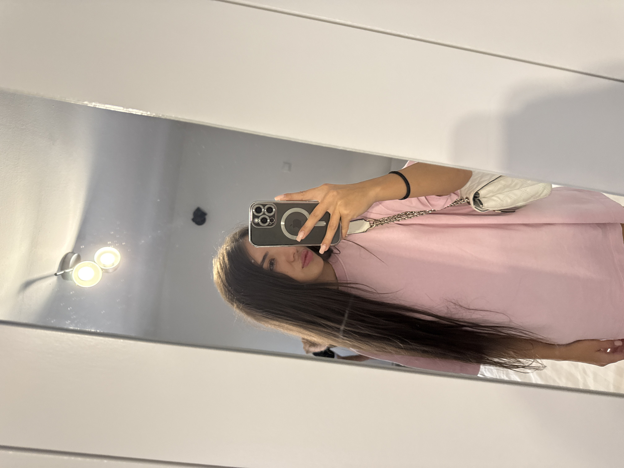 A young woman with long, dark hair is taking a mirror selfie in what appears to be a room with white walls. She is wearing a pale pink oversized t-shirt and has a white quilted purse with a silver chain strap slung over her shoulder. She is holding a dark-colored smartphone in her right hand, which is angled to capture her reflection. The mirror reflects a ceiling light fixture with two circular bulbs. The image does not contain any specific landmarks or visual cues that would place it in Cârțișoara, Romania.