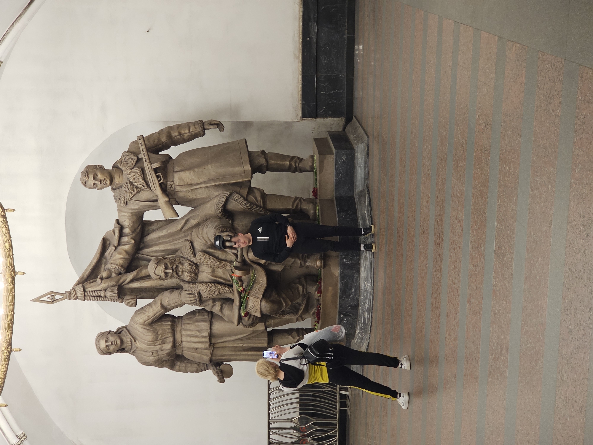 Here is a detailed description of the image:
**Scene:** Indoor, likely a public space such as a metro station or museum.
**Location:** The location is the Heroiv Dnipra metro station, Kyiv, Ukraine.
**Objects:**
* Large, bronze-colored sculpture depicting three figures, possibly soldiers.
* Flag or banner held by one of the figures in the sculpture.
* Red roses placed at the base of the sculpture.
* Marble-like flooring with stripes.
* A black column.
* A decorative railing or barrier in the foreground.
* Smartphone held by a person.
**People:**
* A person in a black tracksuit and black cap is standing in front of the statue.
* Another person is standing and holding a smartphone, possibly taking a picture of the statue or the person standing in front of it. This person is wearing a black tracksuit.
**Actions:**
* The person in black is standing near the statue, likely posing.
* The other person is taking a picture with their smartphone.
**Environment:**
* Walls are light-colored, possibly white.
* The area seems well-lit.
**Interactions:**
* The main interaction is the person taking a photo of the statue with another person posing in front of it.