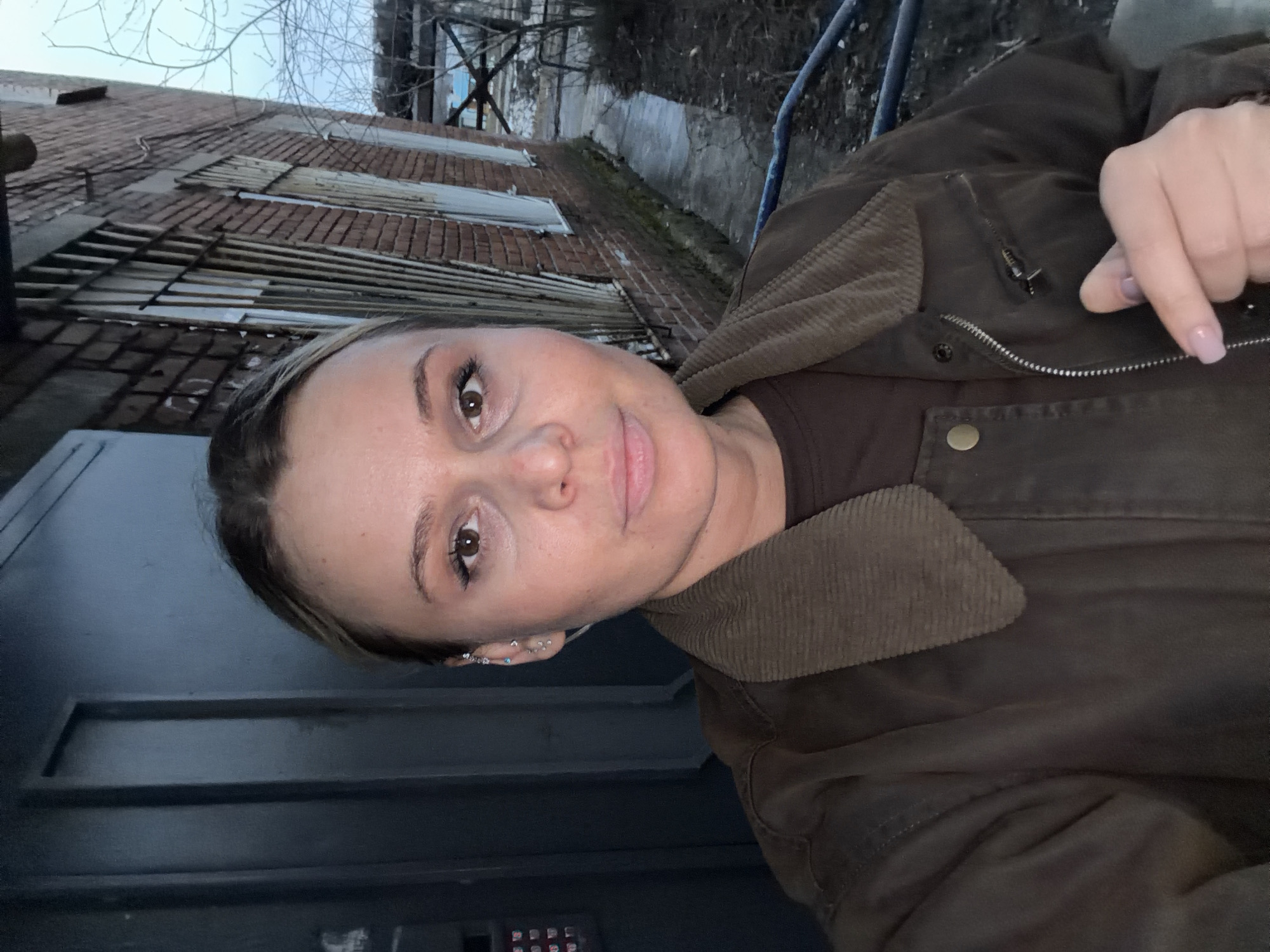 The image is a selfie of a woman wearing a brown jacket with a corduroy collar, taken outside a building in Sarapul, Russia. The building appears to be brick and in a state of disrepair, with boarded-up windows. There is a gray door to the left of the woman with a security keypad. In the background, there is a fence and bare trees, suggesting it may be late fall or winter. The woman has fair skin, brown eyes, and multiple earrings in her left ear. Her fingernails are painted a light purple.