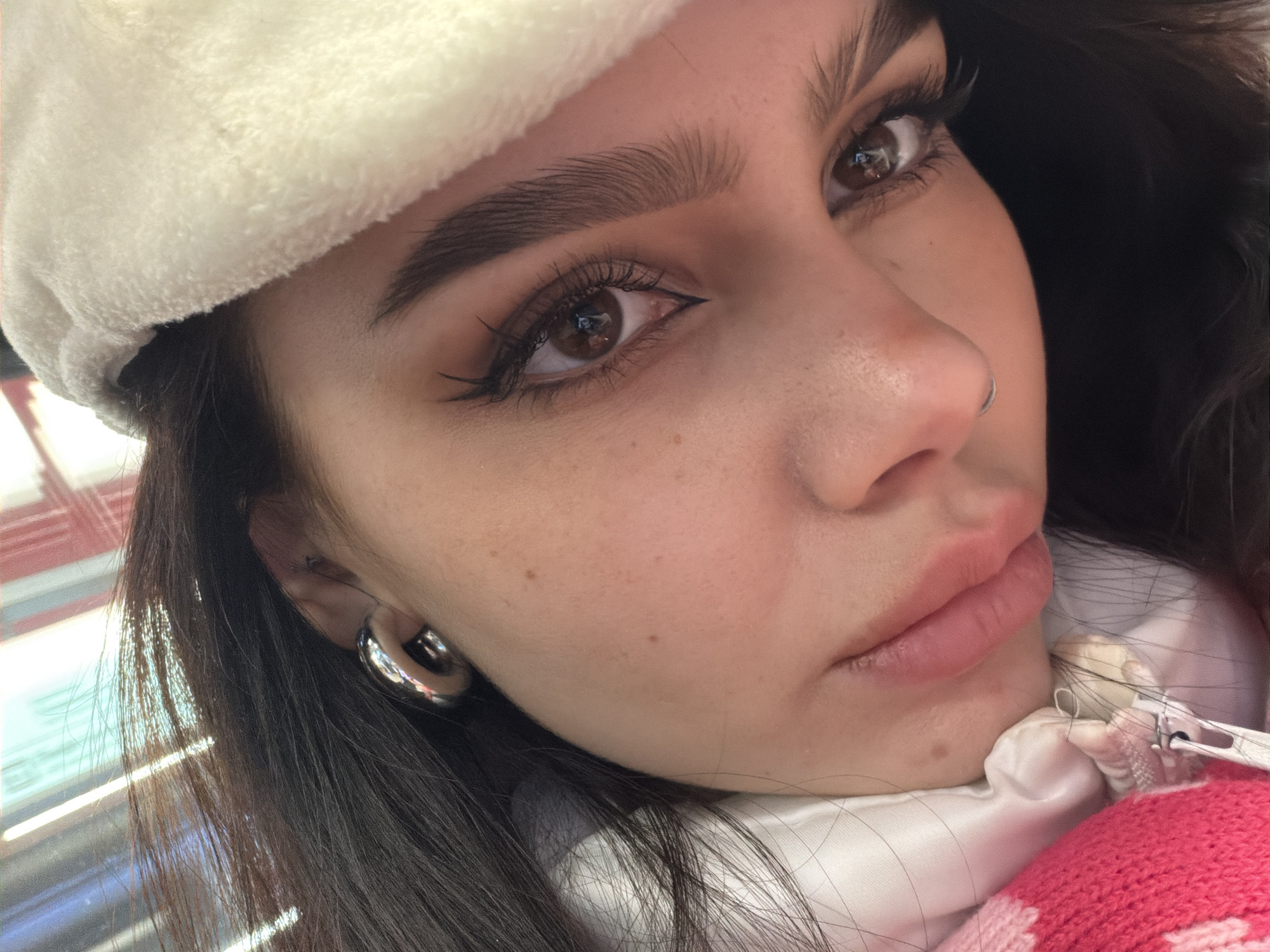 A close-up photograph shows a young woman with dark hair and brown eyes. She is wearing a white, fuzzy hat. She has well-defined eyebrows, eyeliner, and a nose ring. She wears a large, round silver earring. The woman's head is slightly tilted and she appears to be looking into the camera. She is wearing a white jacket with a pink patterned sweater underneath. The location is Moscow, Russia.