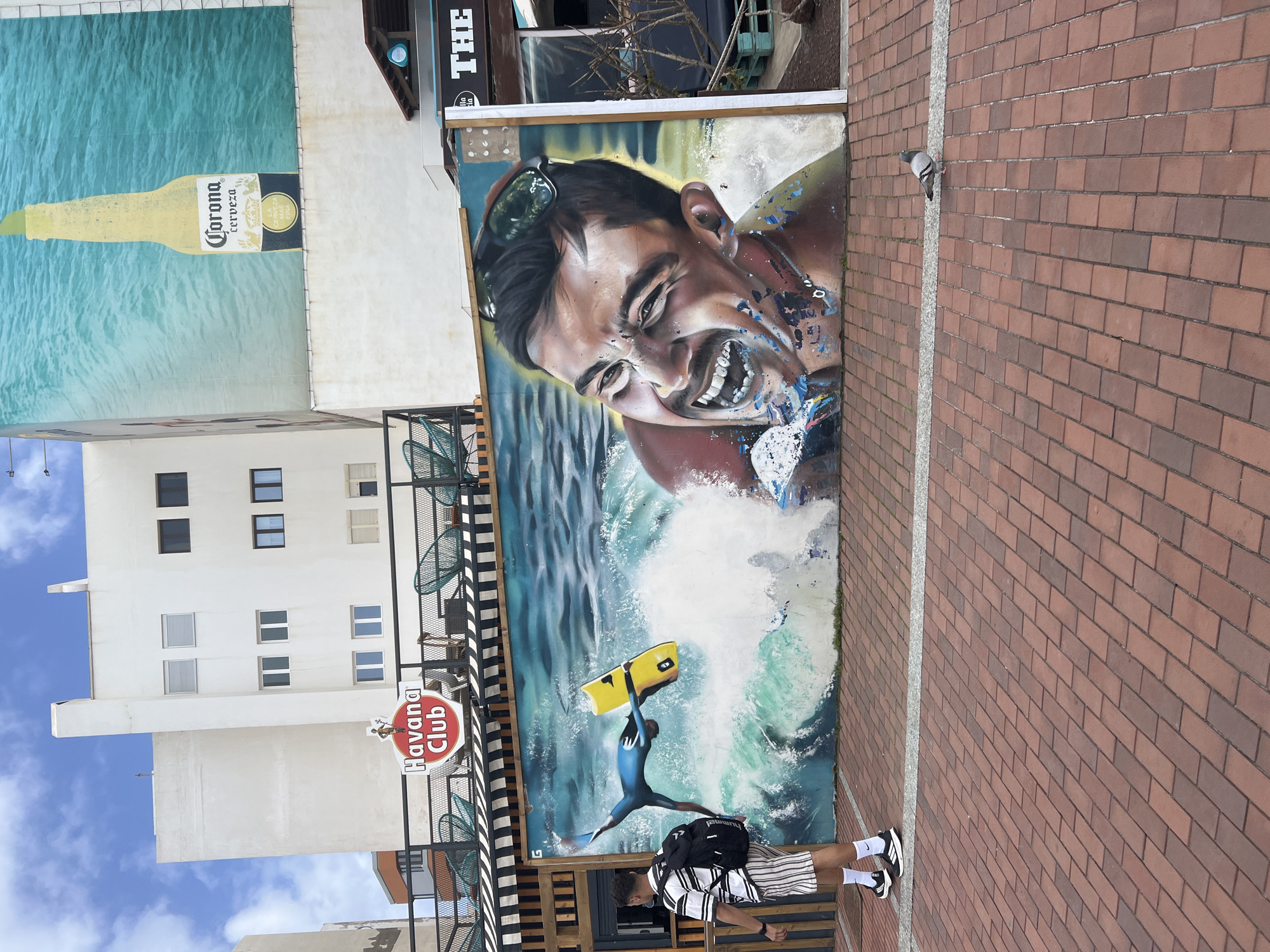 The image shows a street in Las Palmas De Gran Canaria, Spain, featuring several wall murals and two people. On the left is a tall white building with multiple windows and a "Havana Club" sign attached to a black-and-white striped railing. The building's wall is also decorated with a large Corona beer advertisement. Adjacent to this is a wall mural of a person in the water, catching a wave with a yellow surfboard. This mural is adjacent to a wall paved with brown bricks. A pigeon is perched on top of the wall. Standing in front of the mural are two individuals. One of the individuals has a dark backpack on their back. Both individuals are partially obscured by the railing. The environment suggests an urban setting.