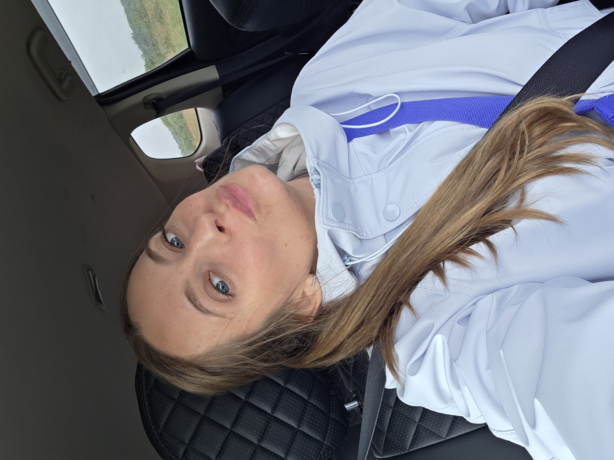 The image displays a woman from an overhead, slightly rotated perspective, likely a selfie taken inside a car. The woman has light skin, striking blue eyes, and long, light brown hair that flows over her shoulder. She is looking directly at the camera with a calm and neutral expression, her lips slightly parted. She wears a light-colored, possibly white or pale blue, hooded jacket with visible buttons and a zipper. A bright purple strap is visible across her chest, under a black seatbelt that diagonally crosses her torso, indicating she is secured in the vehicle.
She is seated in a black car seat featuring a quilted leather design. Through the car's side window and what appears to be a sunroof, a rural landscape unfolds. This external view shows wide, green or brown fields under an overcast, grey sky, suggesting a cloudy day. The setting appears to be a natural, open environment, consistent with the provided location context of Бабстово, Russia. No visible text is present in the image. The scene conveys a sense of a quiet journey or a brief stop during an overcast day.