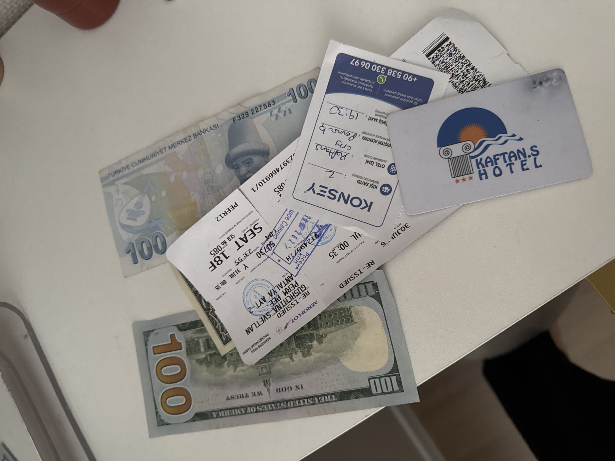 The image displays a close-up, overhead shot of a white, textured surface, likely a table. On the surface, several items are arranged: a portion of a US $100 bill, a Turkish 100 Lira banknote, a boarding pass, and a hotel key card.
The $100 bill is positioned on the left, with the "100" denomination clearly visible. The Turkish Lira banknote is to the right of the US dollar bill, also showing the "100" denomination and Turkish text.
Overlapping these banknotes is a boarding pass. Text on the boarding pass indicates "AEROPLOT," "RE-ISSUED," "GUSHCHIMA," "PERM," and "ANTALYA." The flight details include "SEAT 18F," "Y 11:35," and "30-JUL-23." There is also a stamp with Russian text and what appears to be a date of "01.08.2023." A barcode is also visible on a separate ticket underneath the boarding pass.
A hotel key card, featuring the logo "KAFTAN'S HOTEL," is placed on top of the boarding pass. The card displays the text "KONSEY," "KİŞİ SAYISI: 2," and "VÜSTERİ ACENTASI: cph." The check-in time is listed as "19:30." A phone number, "+90 538 330 06 97," is also printed on the card, along with some text in Russian.
The lighting suggests a well-lit indoor environment. The focus is on the items on the surface, with the background slightly blurred. The scene suggests recent travel or preparation for travel, with the presence of currency, a boarding pass, and a hotel key card. The location of Perm, Russia, is indicated on the boarding pass.