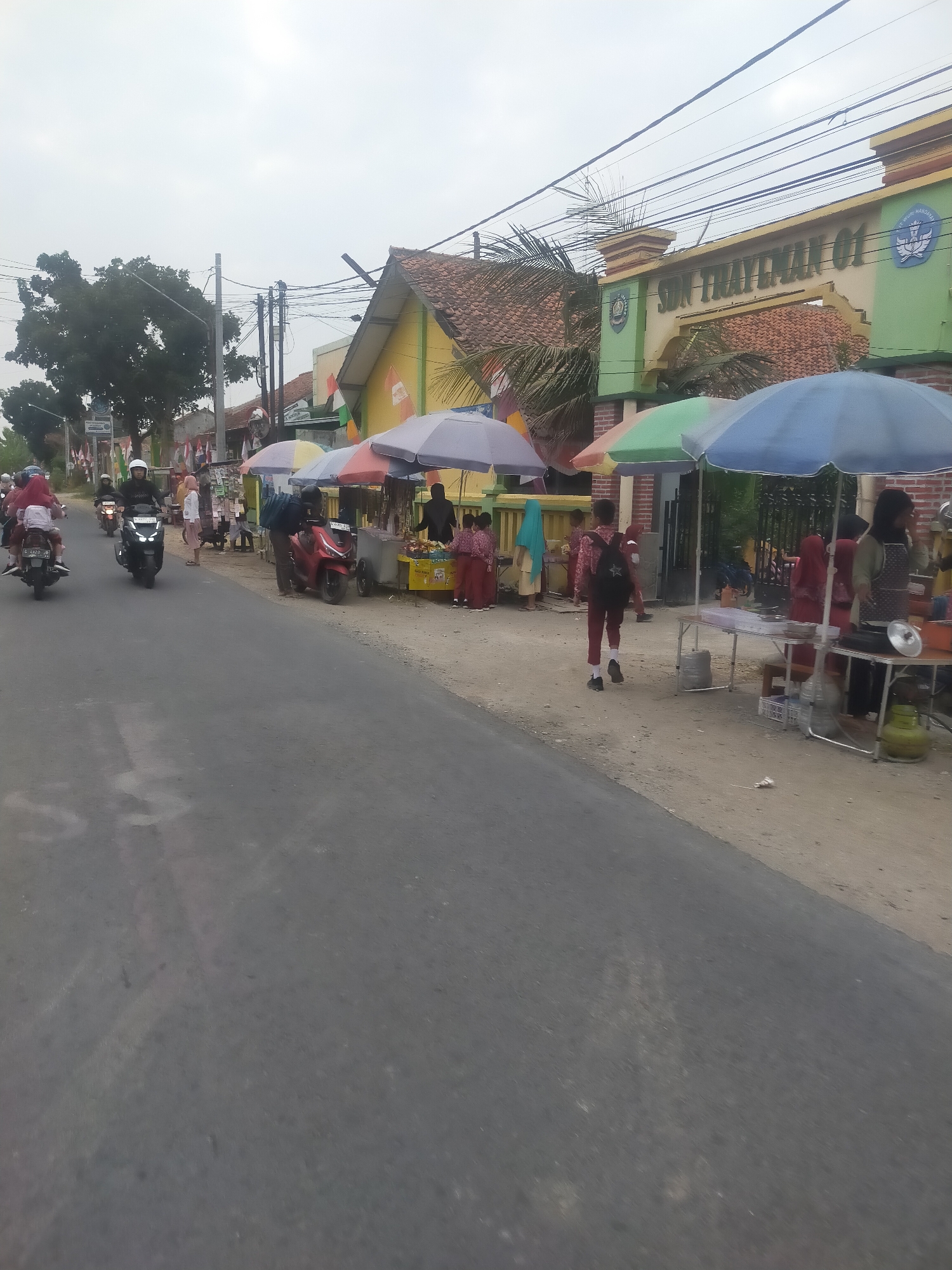 This image captures a scene on a street in front of SDN Trayeman 01 in Trayeman, Indonesia. The street is paved and appears to be two-lane, with a dirt shoulder on the right side. Several people are present, mostly dressed in school uniforms, suggesting it might be a school dismissal or a time when students are arriving or leaving.
On the road, there are several motorcycles. One motorcycle in the foreground on the left has two people on it, with the rear passenger wearing a red jacket. Another motorcycle, black and with a rider wearing a white helmet, is in the middle ground. Further back, another motorcycle is visible.
Along the side of the road, there are several stalls set up under colorful umbrellas, selling various goods, possibly snacks or drinks. One stall has a red scooter parked next to it. People are gathered around these stalls, interacting with vendors. Some individuals appear to be children in school uniforms, and adults are also present, some behind the stalls as vendors, others as customers.
The building directly behind the stalls is the SDN Trayeman 01 school, identifiable by its signage. The school building has a yellow facade and a tiled roof. Flags are displayed along the side of the street, and overhead, power lines crisscross the sky. The overall atmosphere suggests a busy, everyday scene in front of a school, with commercial activity happening on the roadside. The sky is overcast, indicating potentially cloudy weather.
