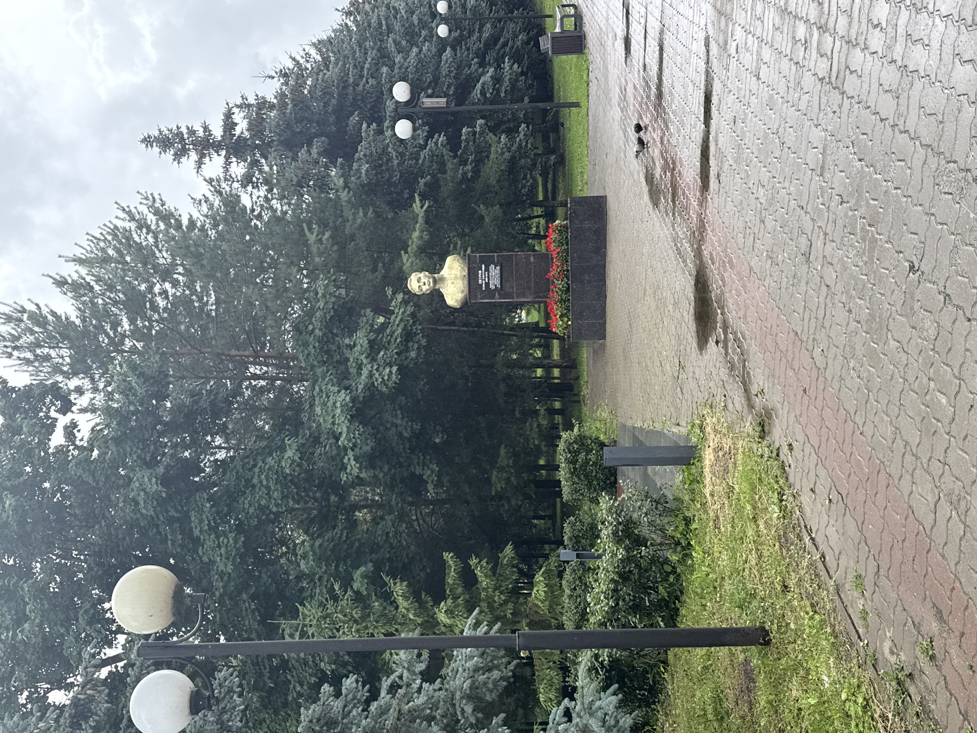 The image depicts a park scene in Kazan, Russia, featuring a monument with a bust of a man. The bust is gold-colored and stands on a dark granite pedestal adorned with red flowers. The pedestal has an inscription in Russian, which reads "ПЕТРОВ ПОРОХОВИЧ" and other text below it. To the right of the monument, a paved walkway with red and grey interlocking bricks is visible, showing puddles from recent rain. Two pigeons are present on the wet pavement. Several lampposts with spherical white lights line the walkway. In the background, dense evergreen trees form a backdrop. To the far right, a park bench is situated. The overall environment suggests a public park or square.