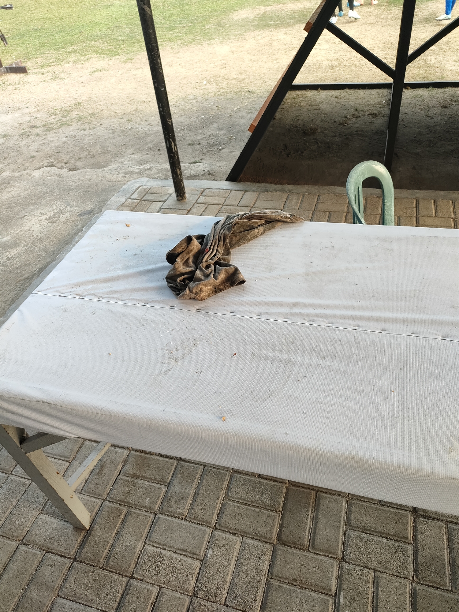 In Banguntapan, Indonesia, a white tablecloth covers a table outdoors. A crumpled brown t-shirt is placed on the tablecloth. Behind the table, a light blue plastic chair is visible, and further back, a wooden structure with dark metal supports stands against a grassy background. The ground is paved with grey interlocking bricks.