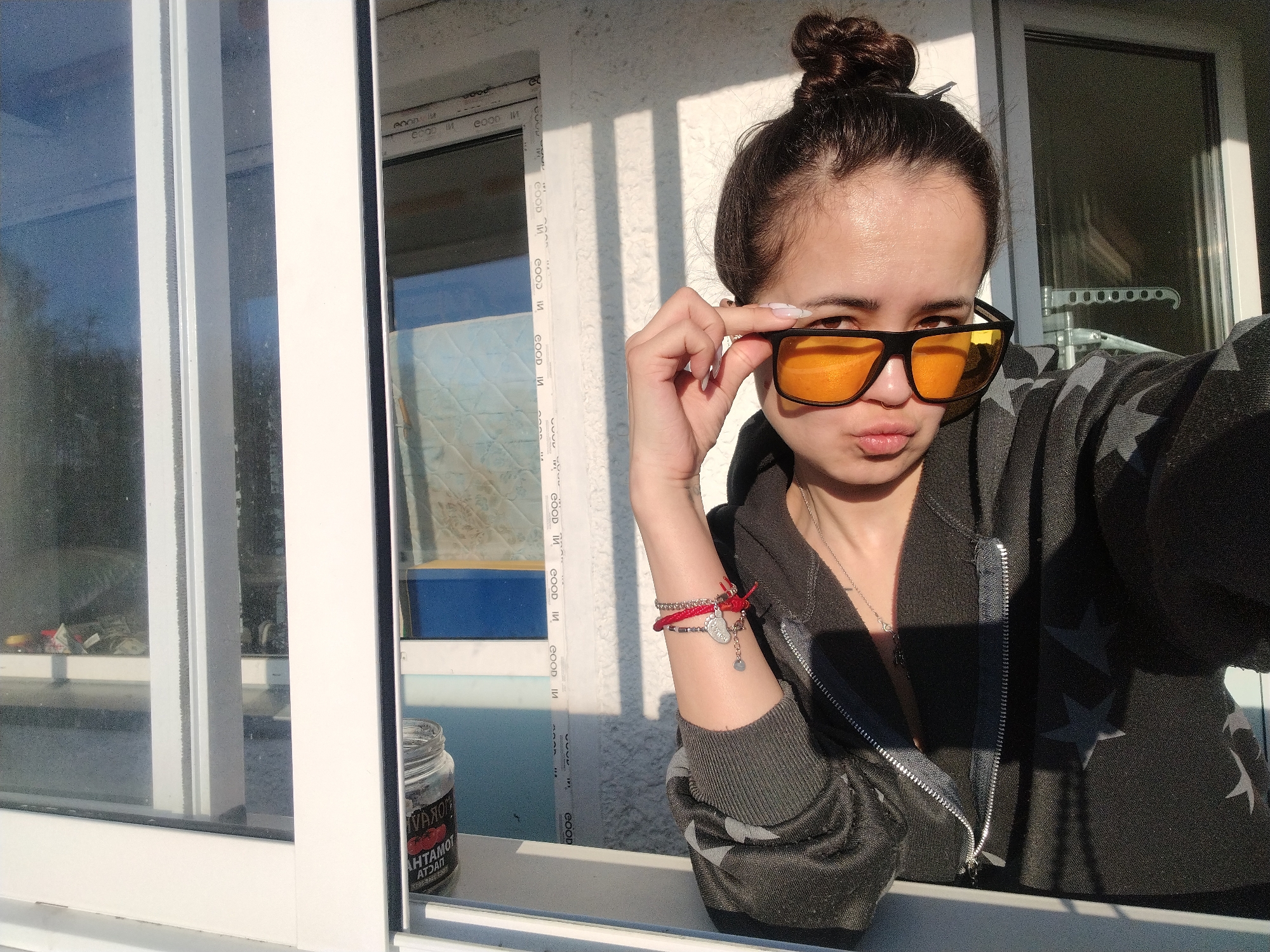 A young woman stands in a window frame. She has dark hair pulled into a bun and is wearing black sunglasses with orange lenses. She is touching the sunglasses with her left hand and making a kissing face. She has a silver necklace and bracelets on her left wrist. She is wearing a gray zip-up hoodie with stars on it. The window frame is white, and there is a jar on the windowsill. The OCR suggests that the inscription on the jar contains the words "VAROM", "AHTAMOT", and "ATDAN". Outside the window, there is a view of a building and trees. The OCR also indicates the presence of the inscription "GOOD IN" multiple times, suggesting that this might be a branding or a printed mark on the window frame itself. There is no information in the image to indicate its location in the city of Noviy, Russia.