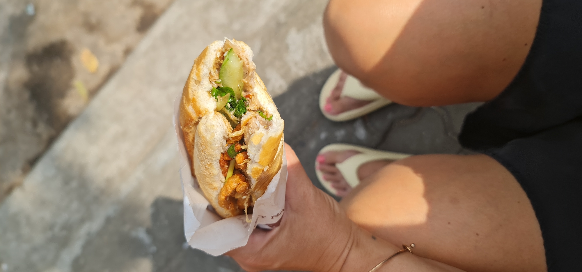 The image shows a person holding a Banh Mi sandwich in an outdoor setting, likely on a street or sidewalk. The sandwich is a Vietnamese baguette, cut open to reveal fillings including sliced meat, cucumber, and green herbs like cilantro. It is partially wrapped in white paper, with the text "NA" visible on the wrapper.
The person's hands hold the sandwich in the foreground. In the background, the person's bare legs are visible, adorned with light-colored flip-flops and pink-painted toenails. The ground beneath them appears to be a concrete or paved surface. Strong, distinct shadows indicate bright daylight and sunny weather. The overall scene suggests a casual meal taken on the go. Given the context, this scene is set in Đà Nẵng, Vietnam.