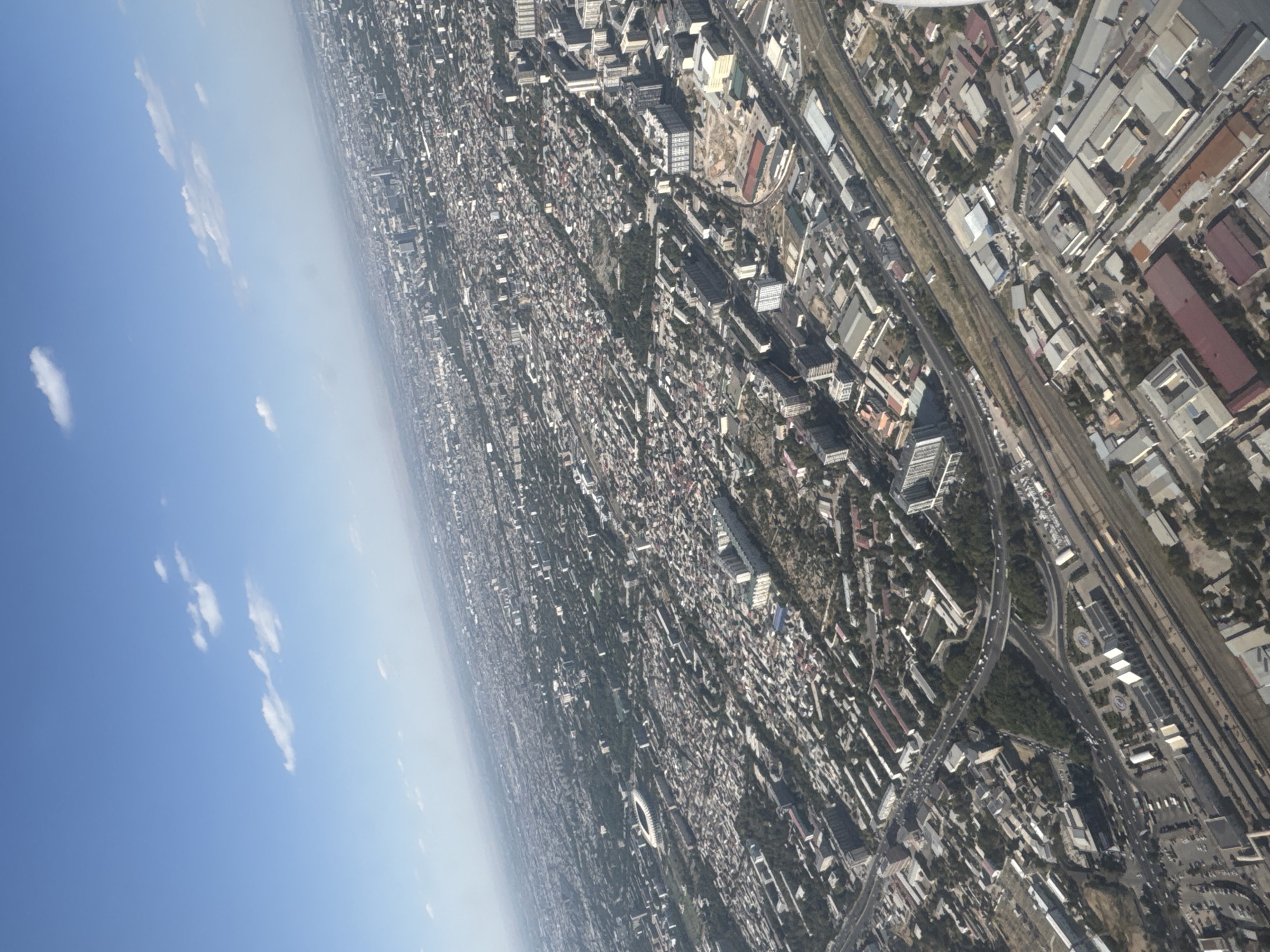 The aerial image provides a wide-angle view of a densely populated urban area, likely Tashkent, Uzbekistan, characterized by a mix of residential buildings, commercial structures, and extensive infrastructure. The scene is dominated by a sprawling cityscape under a clear blue sky with scattered white clouds.
In the foreground, the image captures a complex network of roads, including a highway with moderate traffic, and railway lines. Adjacent to these transportation arteries are numerous industrial or commercial buildings with varied roof types, many appearing to be warehouses or factories. Some of these structures feature large, rectangular footprints and metallic roofing. Parking lots with visible vehicles are also present in this area.
Moving further into the mid-ground and background, the urban landscape transitions to predominantly residential zones. These areas are marked by a dense arrangement of apartment blocks, ranging in height and architectural style. Many of these buildings are long and rectangular, typical of Soviet-era urban planning. Between these structures, green spaces, parks, and tree-lined streets are interspersed, suggesting a city with significant tree cover. A prominent circular stadium, likely a sports venue, is visible in the mid-ground left. The horizon shows a hazy expanse of the city fading into the distance, with the subtle outline of hills or mountains in the far background, indicating the geographical context of the city.
The overall impression is that of a large, established city with a functional, albeit somewhat dated, urban fabric. The image does not clearly depict any individuals or specific human interactions, focusing instead on the built environment and its organization. The lighting suggests it is daytime, with the sun casting shadows, though these are not pronounced due to the aerial perspective and angle. The image is captured from a high vantage point, suggesting it was taken from an aircraft or a very tall structure.