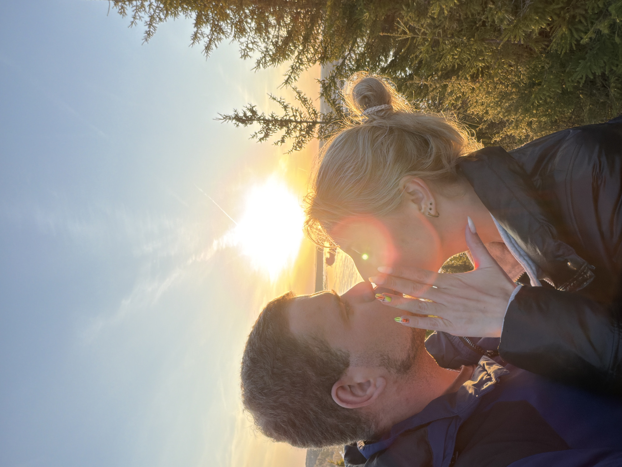 In a landscape setting, a man and a woman are in a close, intimate embrace, their faces near as if they are about to kiss or are kissing. The woman has blonde hair pulled back into a bun and is wearing a dark jacket. Her nails are painted with a bright, abstract design. The man, with short graying hair and stubble, is wearing a dark jacket with a blue underlayer. The sun is low in the sky, creating a warm, bright light and flare. The background includes trees and possibly a body of water. The location may be in the city of Ольховка, Russia, as specified.