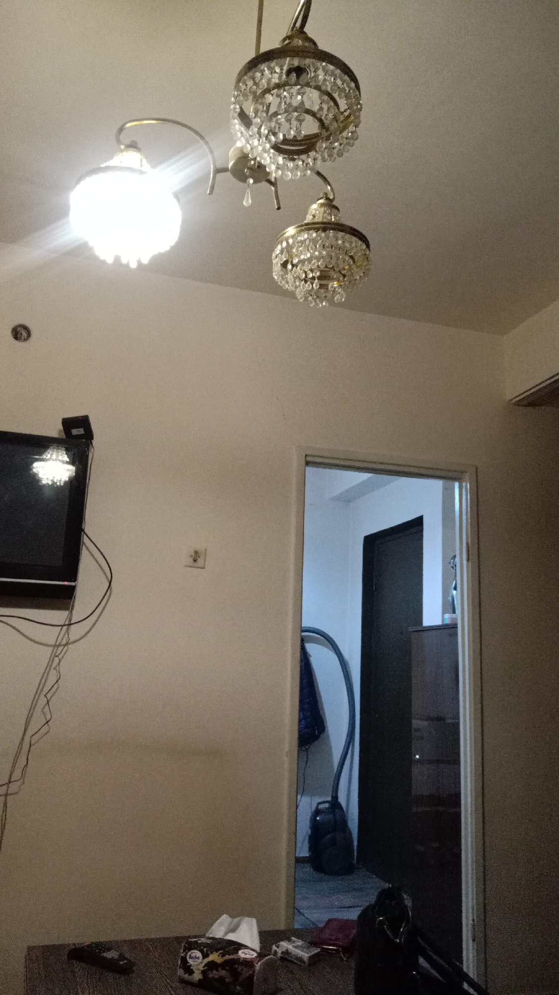 The image is an interior shot of a room with white walls and a white ceiling. A three-light chandelier hangs from the ceiling. A television is mounted on the left wall, with electrical wires hanging down. Below the television, on the wall, is a light switch. A doorway is visible on the right, leading into another room where a vacuum cleaner and a dark cabinet are seen. In the foreground, on a table, there are some objects including a remote control, a tissue box, and other items. The image does not contain any indicators that would identify the location as Yerevan, Armenia.