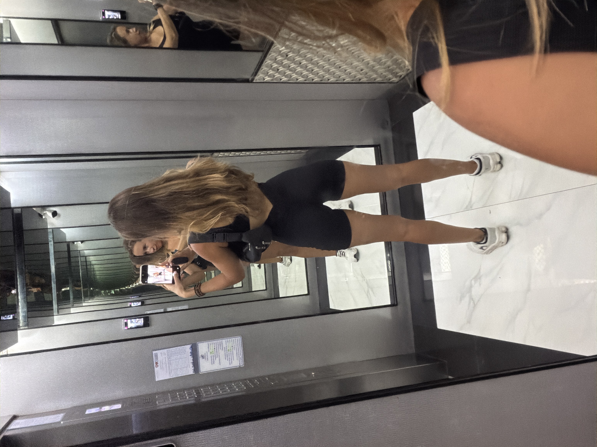 A young woman in a black exercise outfit is standing inside an elevator with mirrored walls in Pattaya City, Thailand. She is taking a selfie with her phone. The elevator has a white marble floor and gray walls.