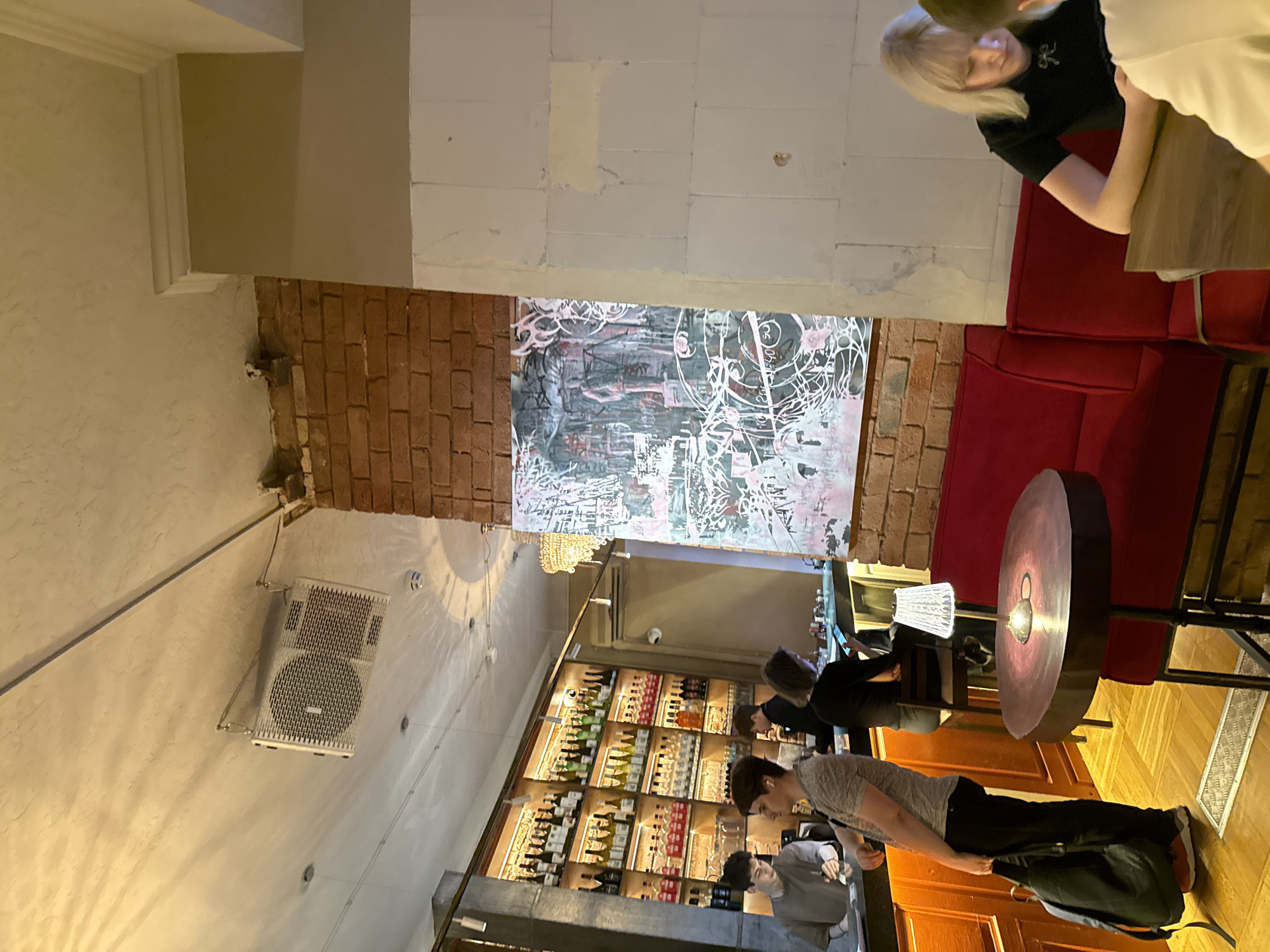 The image shows the interior of a bar or restaurant in Moscow, Russia.
In the foreground and to the right are several people seated at a round, brown wooden table with a metal base. One person wears a black t-shirt and is sitting on a red velvet booth. Another person in a light-colored top appears to be placing a black backpack down.
Behind them, the bar area features a large shelf stocked with numerous bottles of alcohol. Several other patrons are present at the bar, with one wearing a black shirt.
Above the bar, there's a large, abstract piece of artwork framed by exposed brick. A decorative chandelier hangs above the artwork.
The walls are a mix of exposed brick and painted surfaces, and an air conditioning unit is mounted on the wall. The lighting appears to be dim and atmospheric.