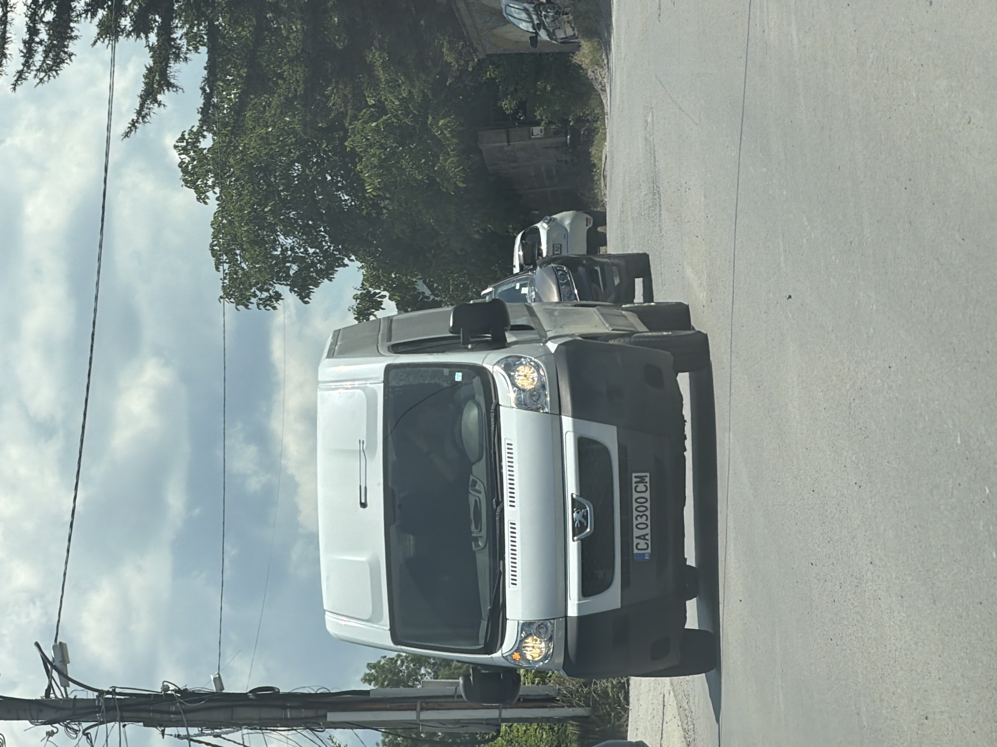 This media file shows a white van with a license plate that reads "CA 0300 CM". The van is parked on a paved road, and there are trees and power lines in the background. The sky is cloudy, suggesting it might be daytime. The van appears to be a delivery vehicle or a personal utility vehicle. There are no people visible in the image. The overall scene suggests a quiet street, possibly in a suburban or industrial area. The image is oriented vertically, making it a bit disorienting to view. The license plate suggests the vehicle is registered in Bulgaria, and the presence of power lines and trees are common elements in many urban and rural landscapes.
