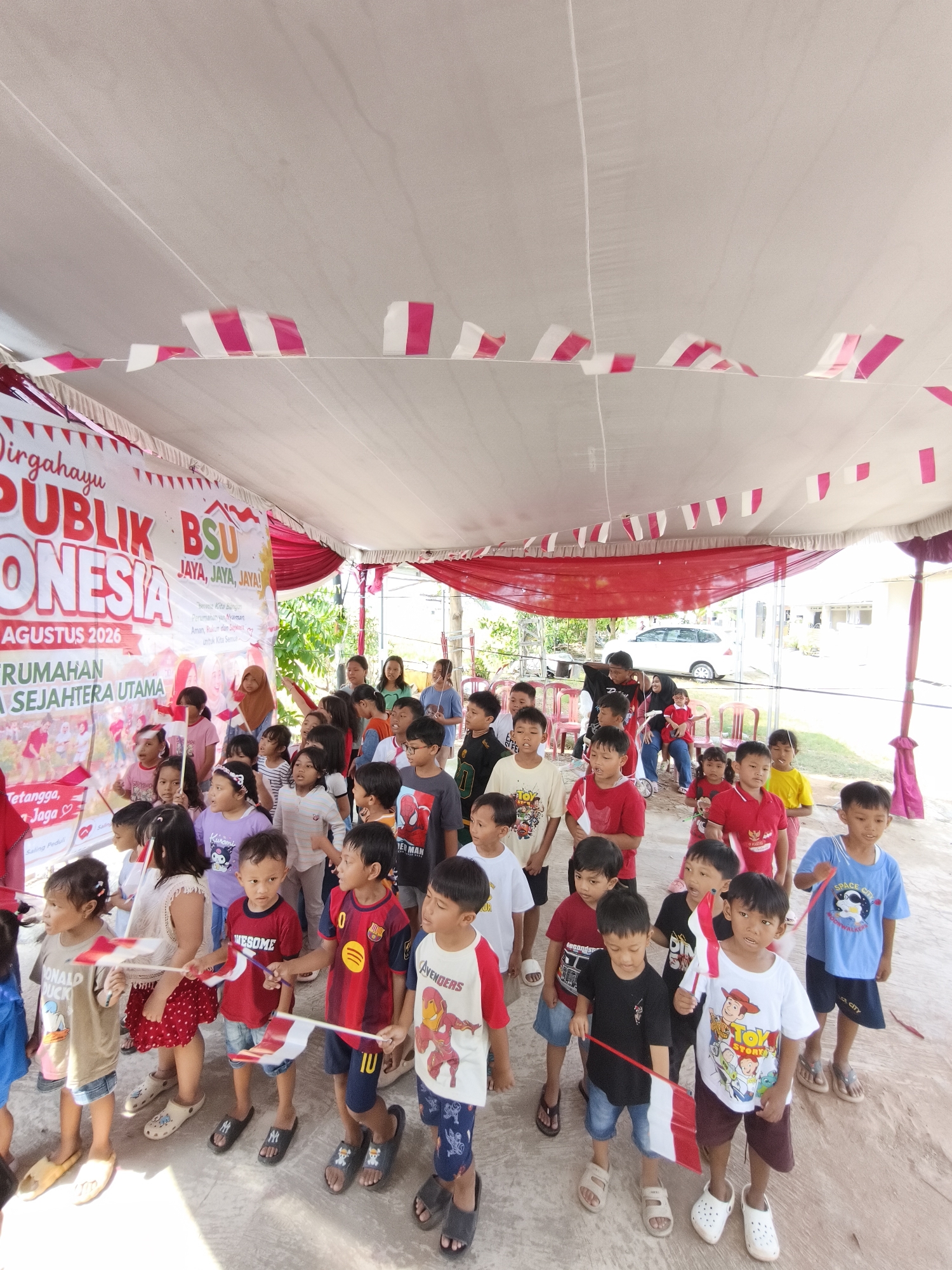 The image captures an outdoor gathering of children and adults under a tent decorated with red and white pennant banners, seemingly for a national celebration. A large banner behind the crowd reads "Dirgahayu PUBLIK INDONESIA" and mentions "AGUSTUS 2026," suggesting an Independence Day or similar patriotic event. The children are holding small Indonesian flags.
The setting appears to be a public space, possibly a community center or an event ground in Bandar Lampung, Indonesia. The ground is paved, and some chairs are visible. In the background, a white car is parked near a building, and lush green foliage can be seen, indicating a tropical environment. The overall atmosphere is festive, with the children appearing engaged and excited.